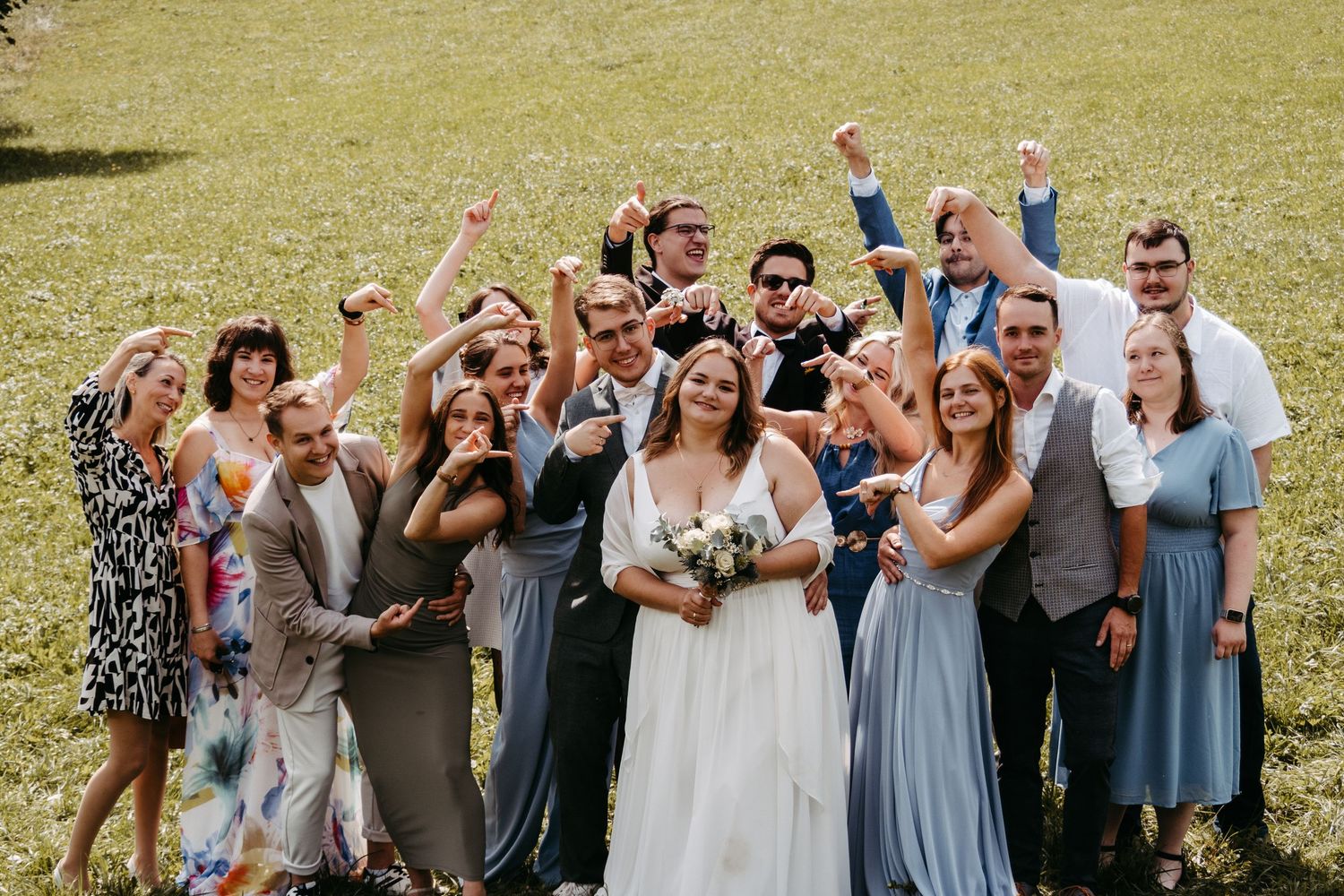 Joyful wedding party celebrating outdoors on green grass, bride in white gown holding bouquet with cheering guests.