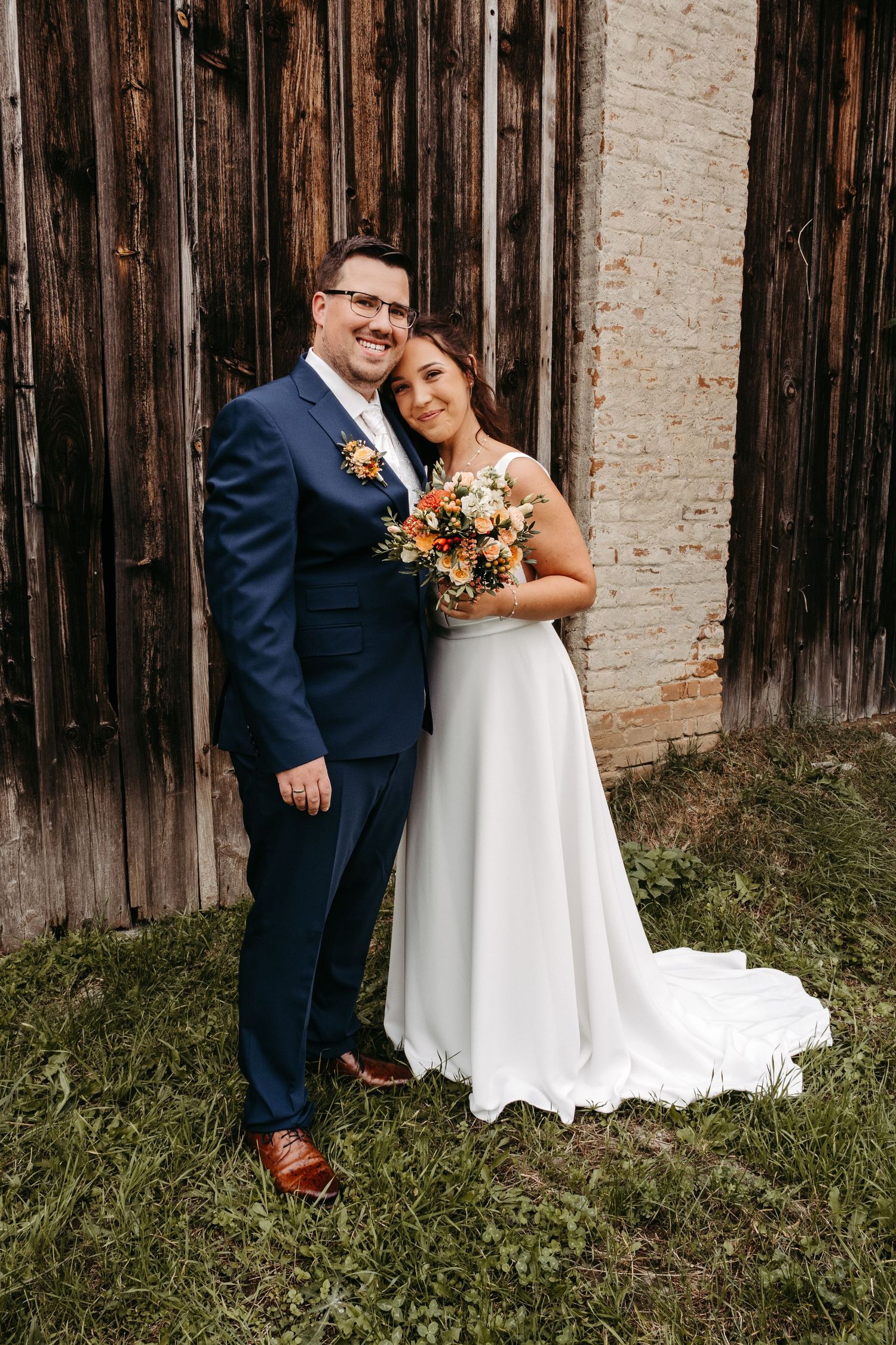 A bride and groom pose together in front of a rustic wooden barn holding an autumn bouquet.