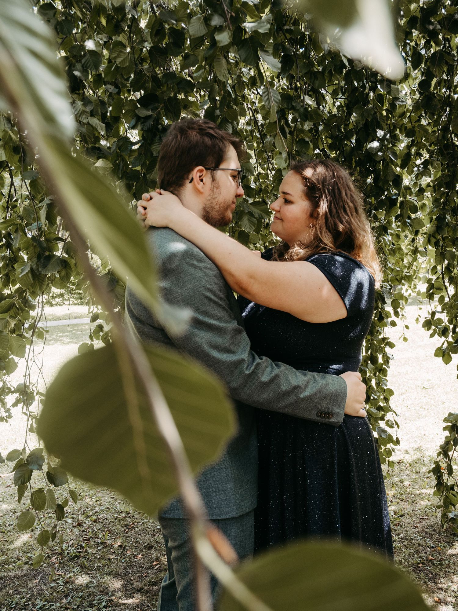 Couple embracing romantically under cascading green leaves in an outdoor garden setting.