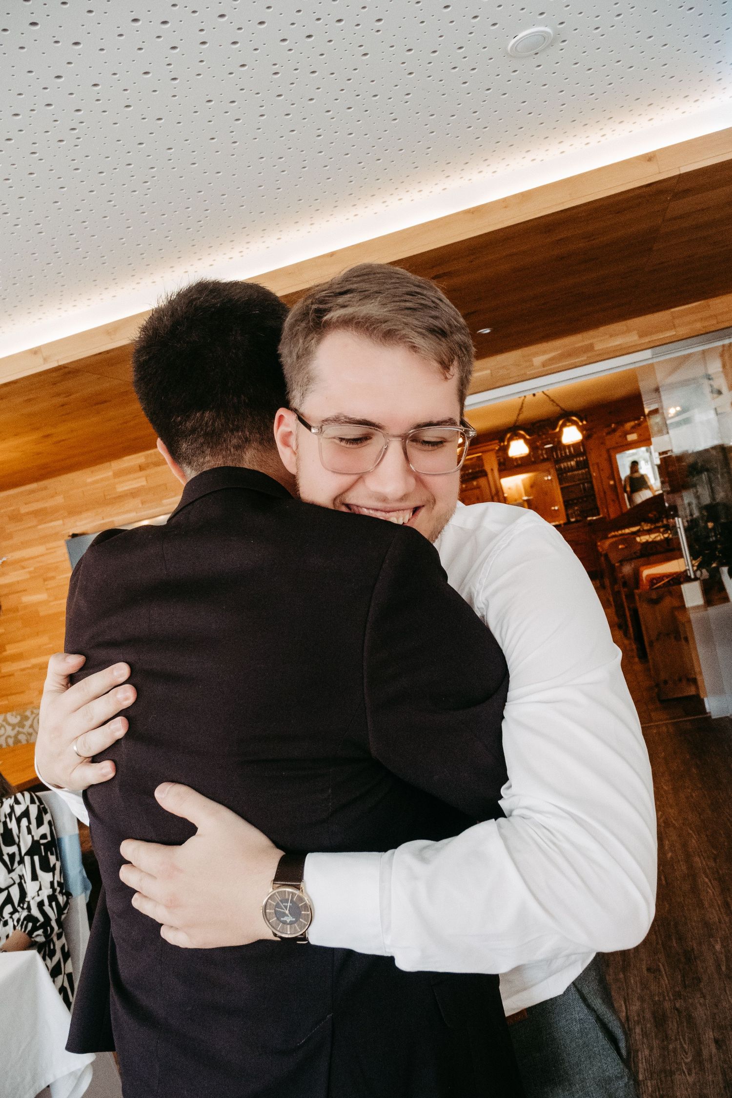 Two men sharing a warm hug in a restaurant, one smiling broadly wearing glasses and a white shirt with a watch.