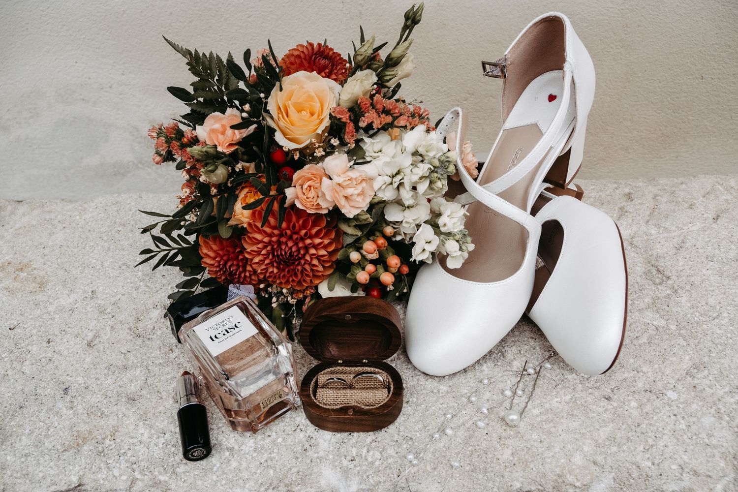 Bridal flat lay featuring white Mary Jane shoes, floral bouquet, perfume, lipstick, and ring box on a marble surface.
