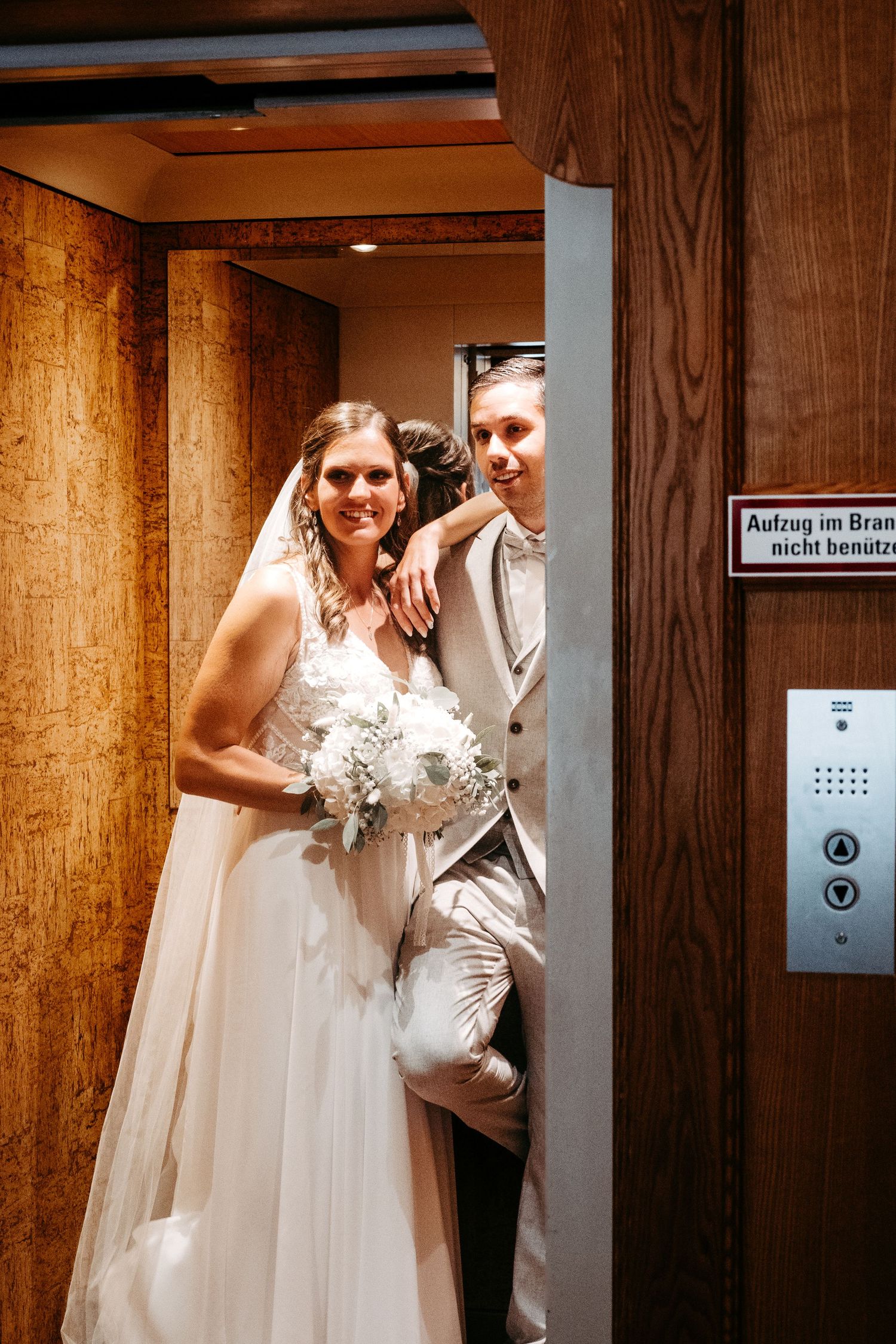 A bride in a white gown holding flowers and groom in gray suit pose together inside a wooden elevator.