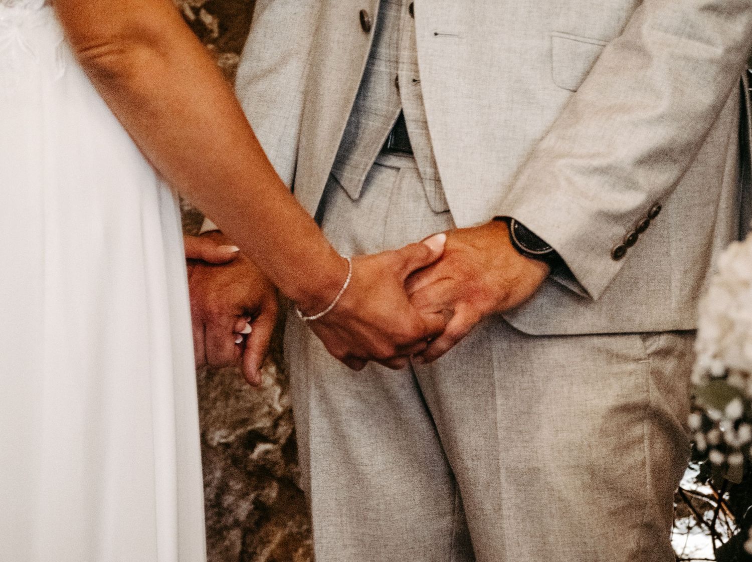 Bride and groom holding hands at wedding ceremony, bride in white dress and groom in light gray suit.