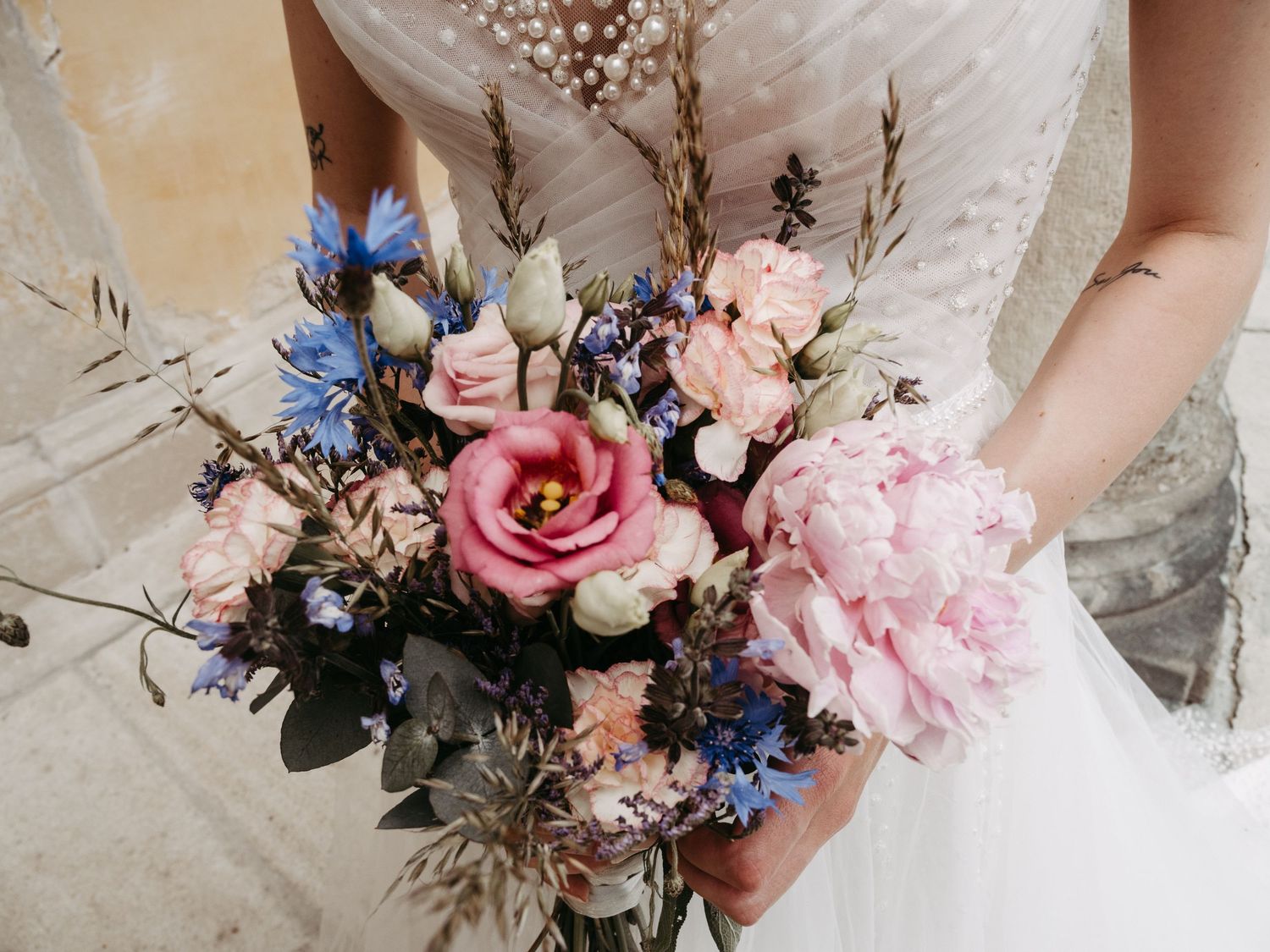 Bride holding a romantic wedding bouquet with pink peonies, blue cornflowers, and wild grasses in a rustic outdoor setting.