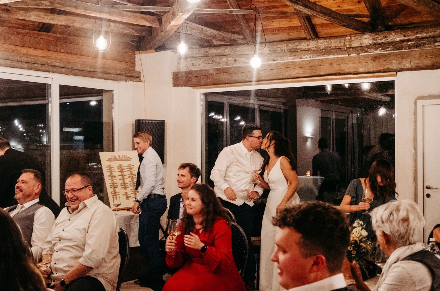 Bride and groom kissing while guests celebrate at lively indoor wedding reception.