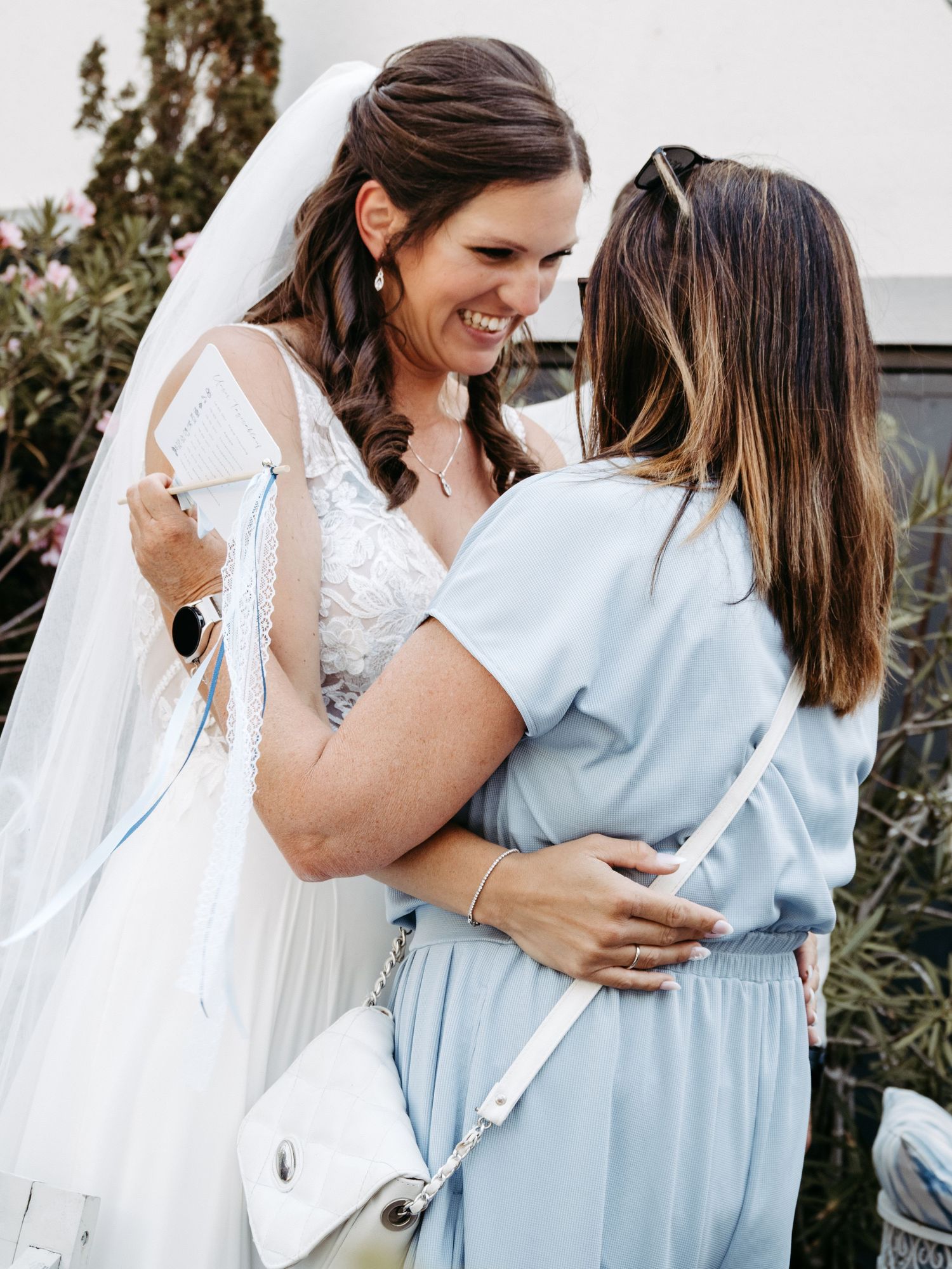 Bride in white lace gown and veil embracing a woman in light blue dress, sharing a joyful moment outdoors.