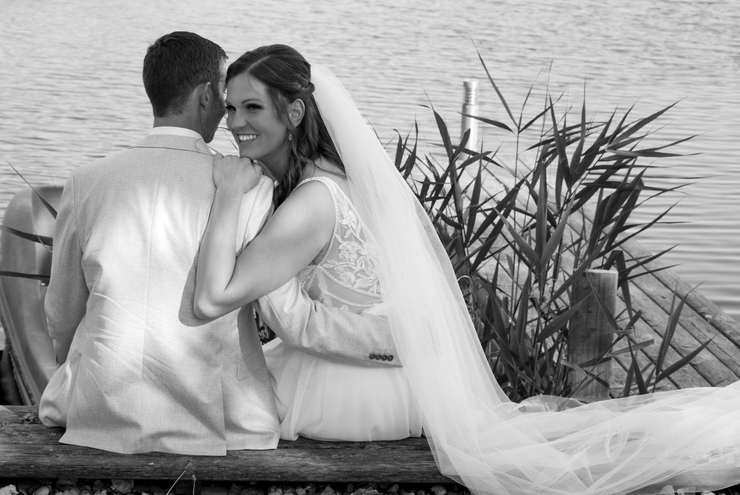 Black and white photo of a bride and groom sitting together by a lake, sharing a romantic moment on their wedding day.