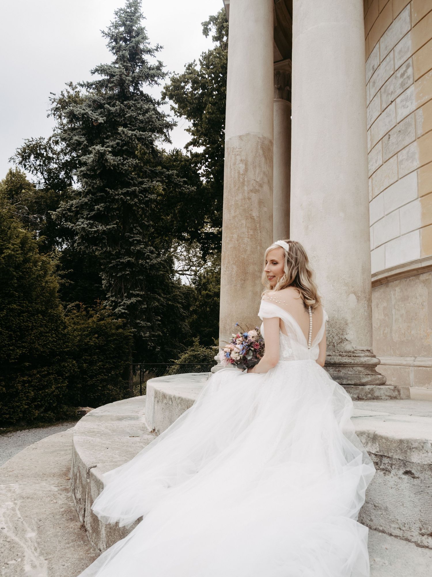 Bride in off-shoulder white tulle gown holding bouquet on stone steps beside classical columns and lush green trees.