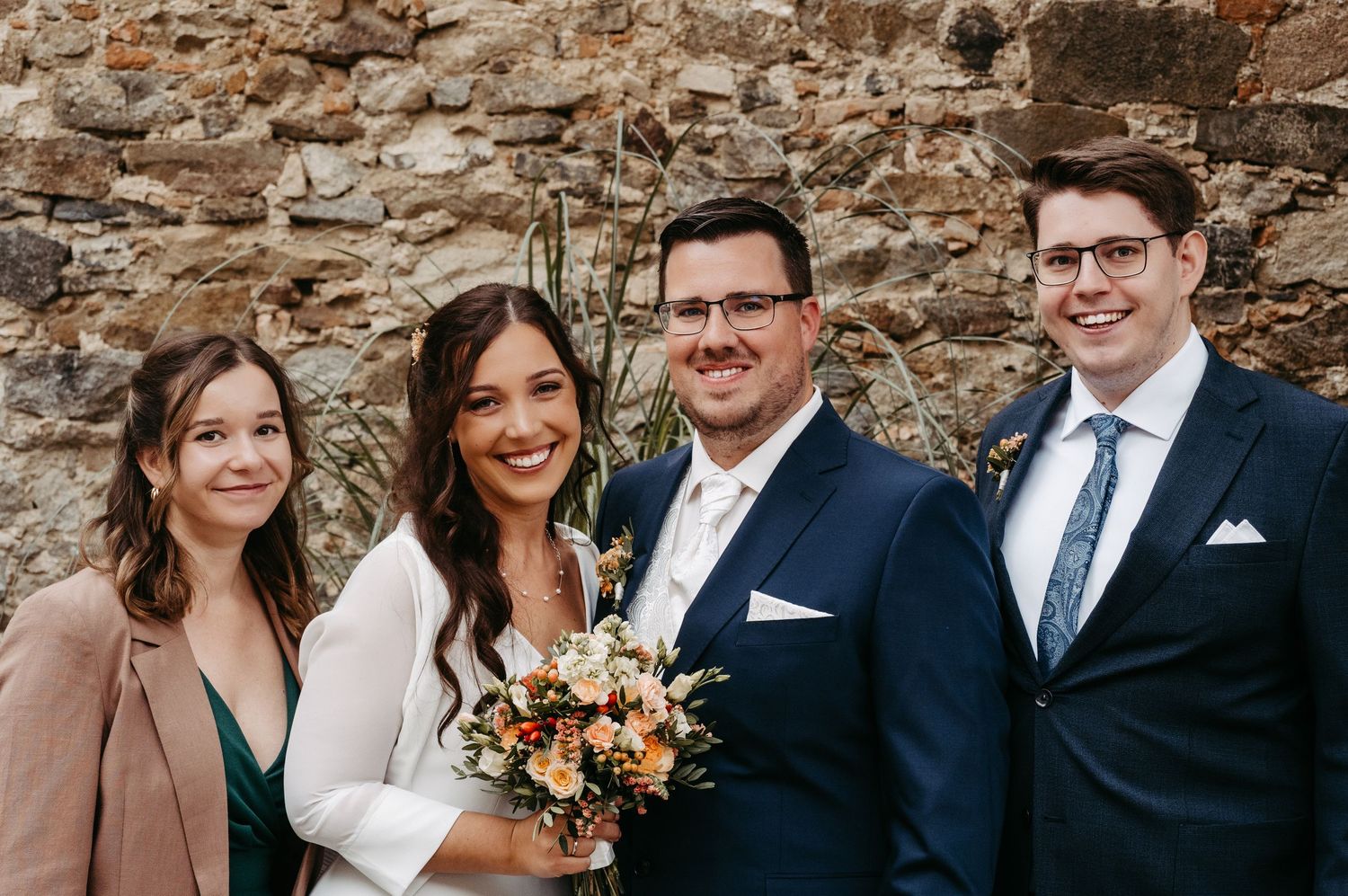 Wedding couple with guests posing outdoors against stone wall, bride holding bouquet, groom in navy suit.