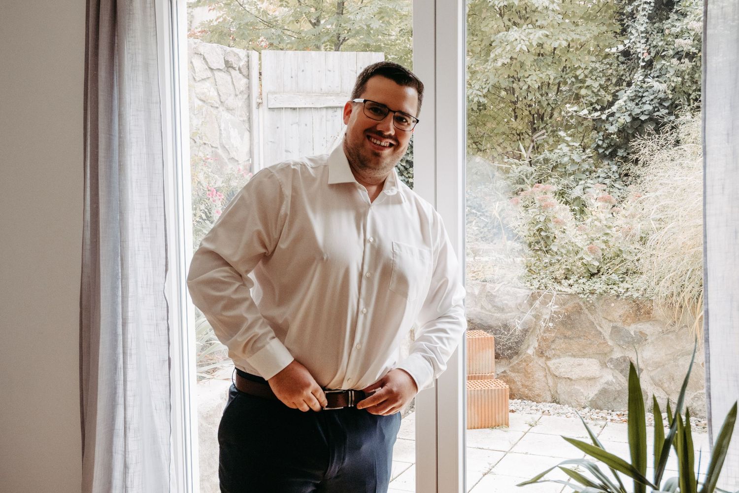 Man in white dress shirt adjusting belt near bright window with garden view, getting ready for formal occasion.