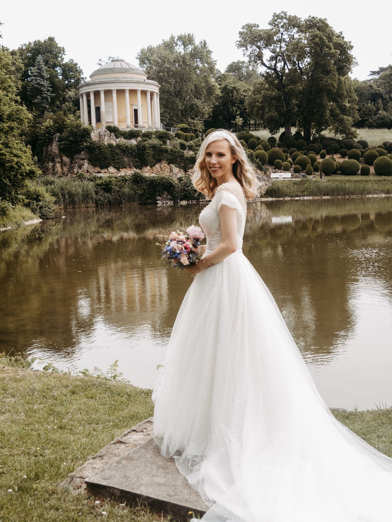 Bride in white ball gown holding bouquet stands by a pond with a classical rotunda and lush gardens in the background.