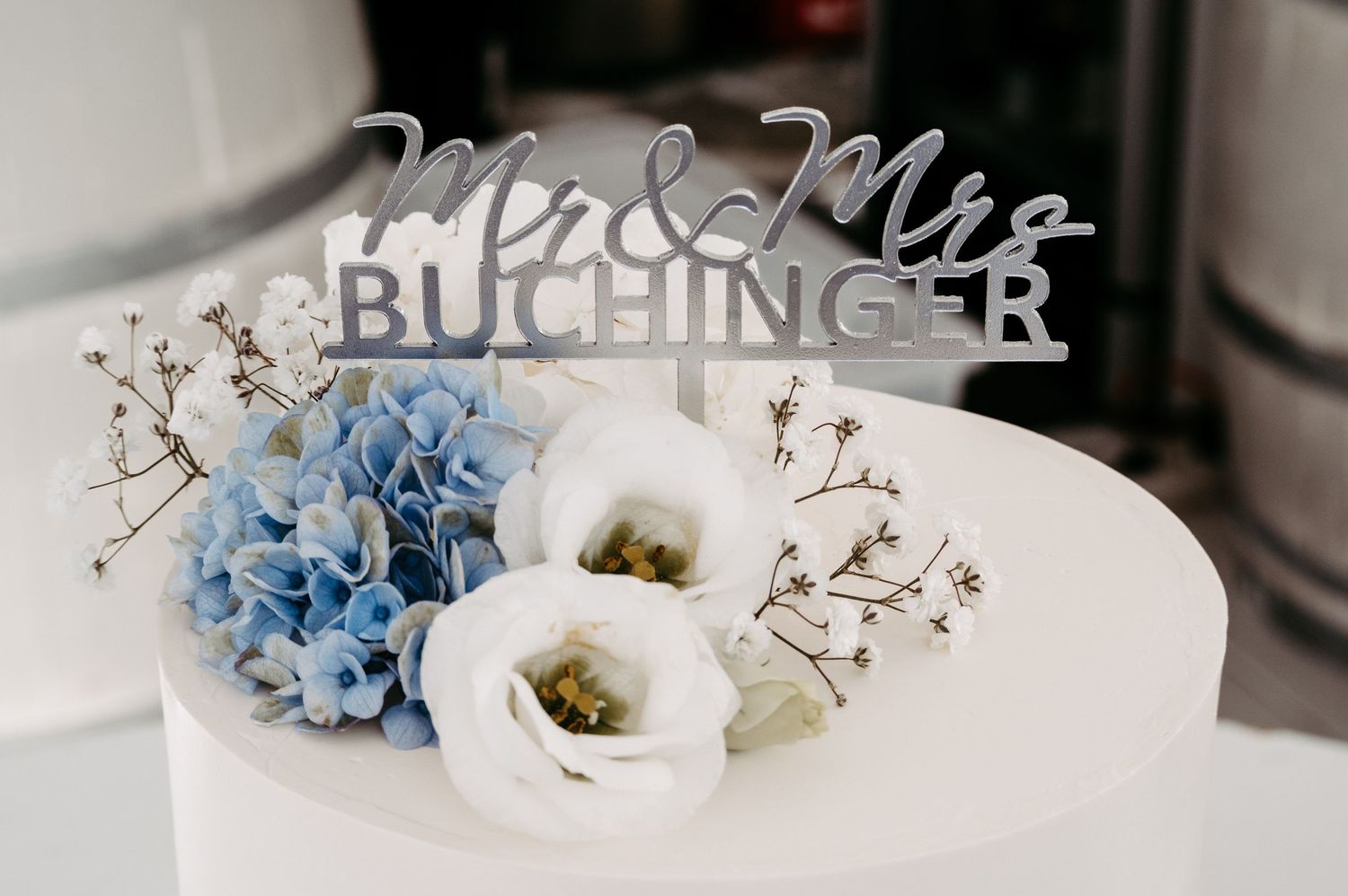 White wedding cake with Mr & Mrs Buchinger silver topper, blue hydrangeas, and white roses floral arrangement.