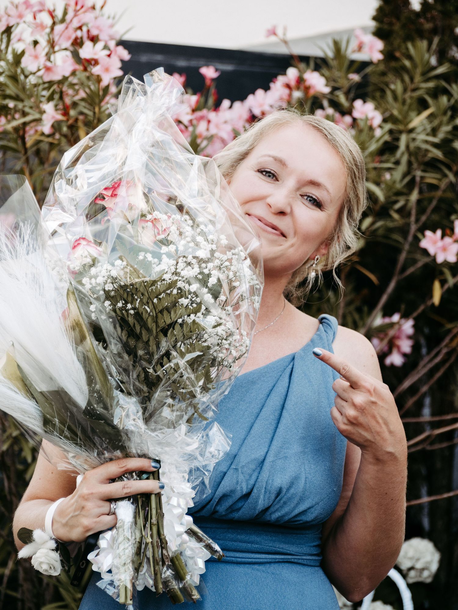 Smiling blonde woman in blue dress holding wrapped flower bouquet outdoors near pink flowering shrubs.