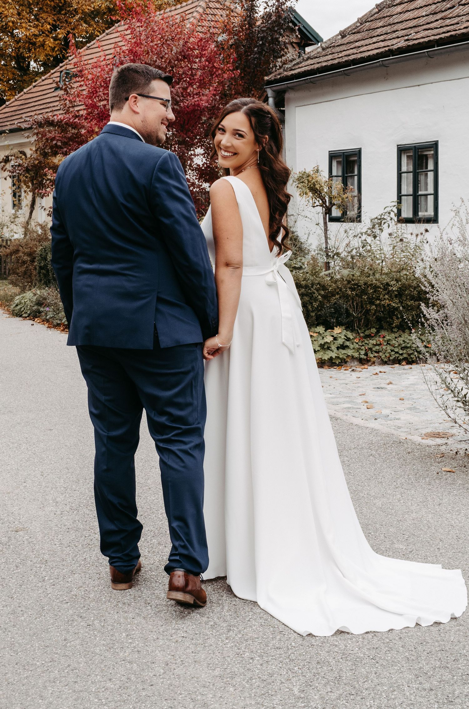 Bride in white gown with bow and groom in navy suit walking together outdoors.