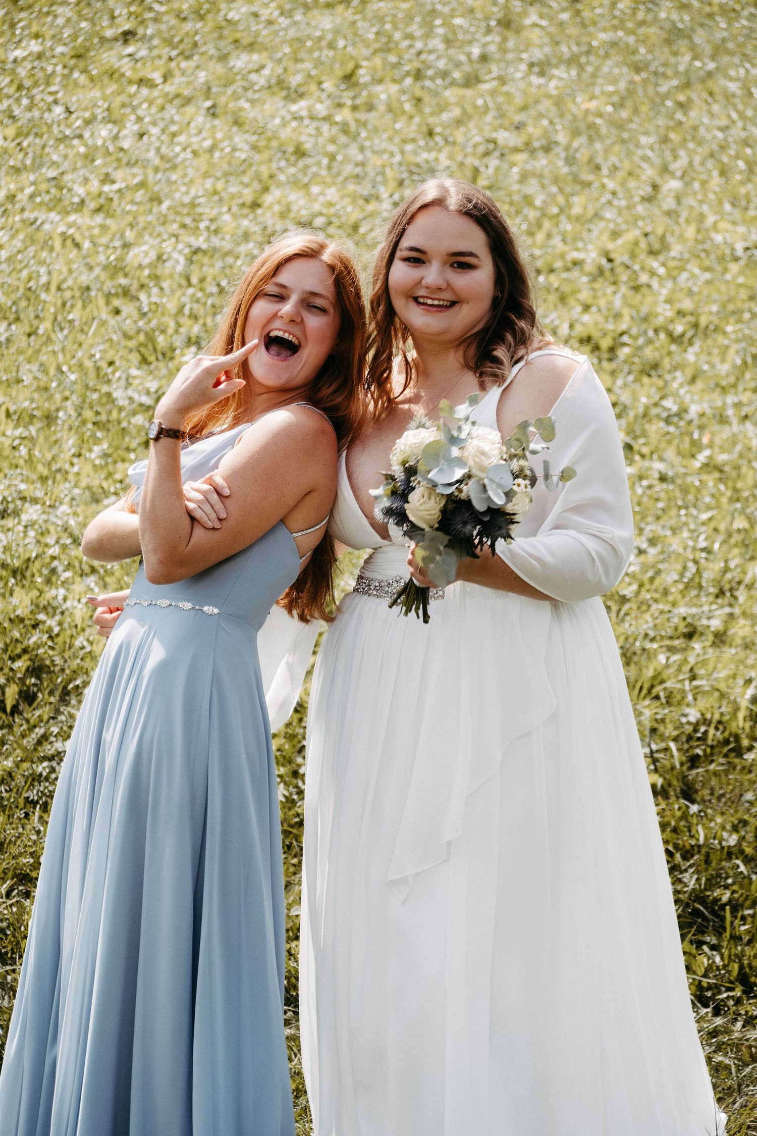 Bride in white gown holding bouquet poses with bridesmaid in blue dress on a sunny day outdoors.