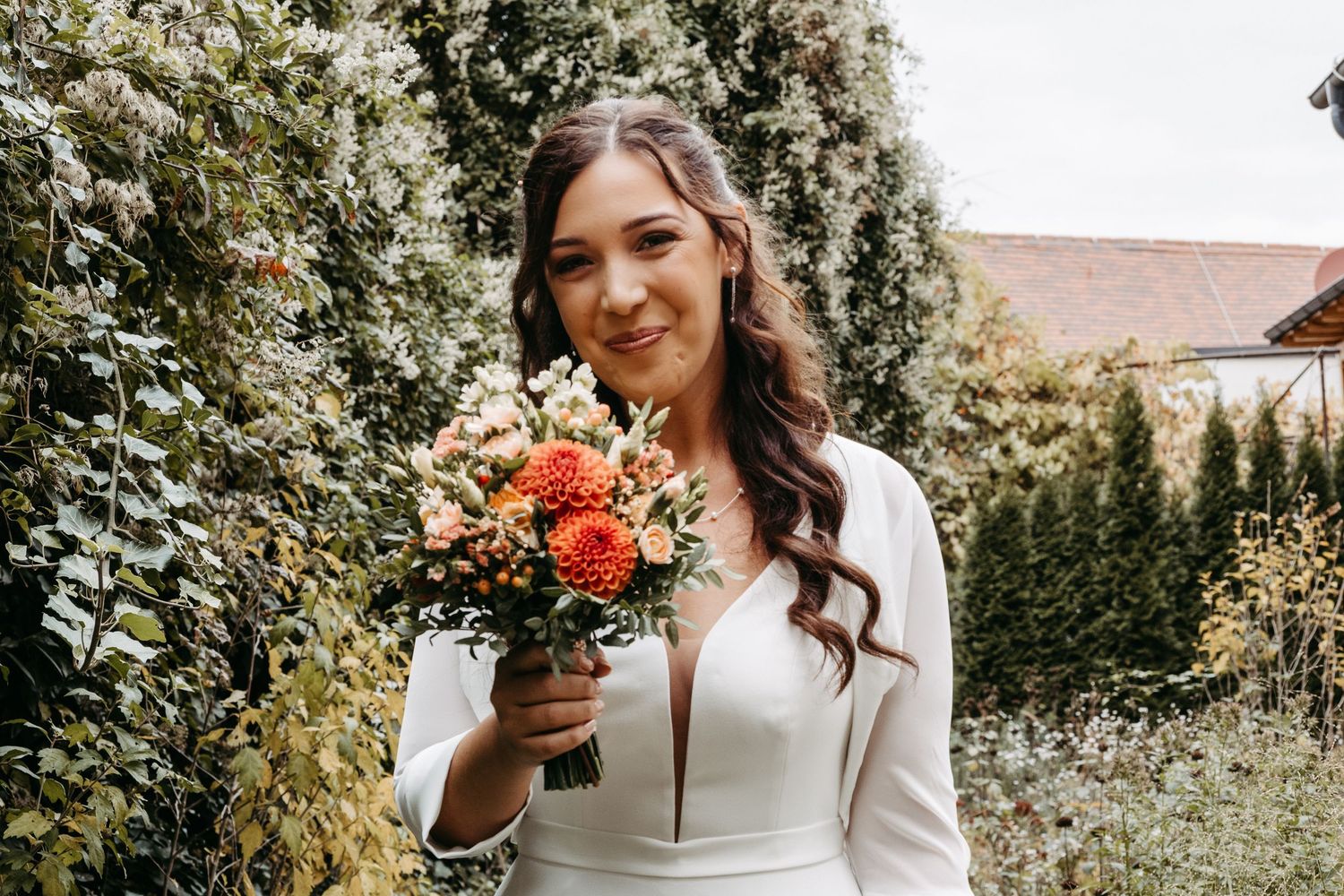 Smiling bride in white dress holding orange and pink bouquet in lush garden setting.