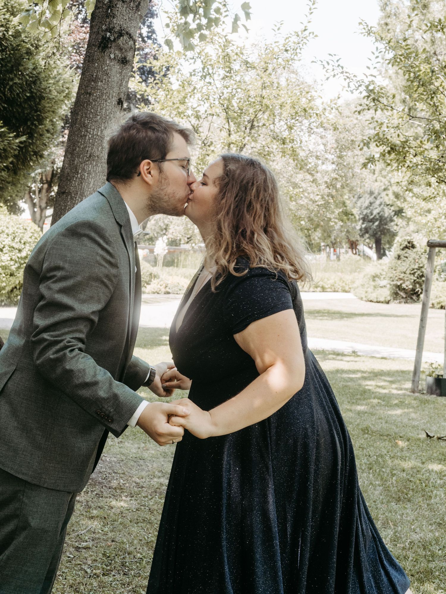 Couple sharing a romantic kiss outdoors in a park, man in gray suit and woman in black dress holding hands.