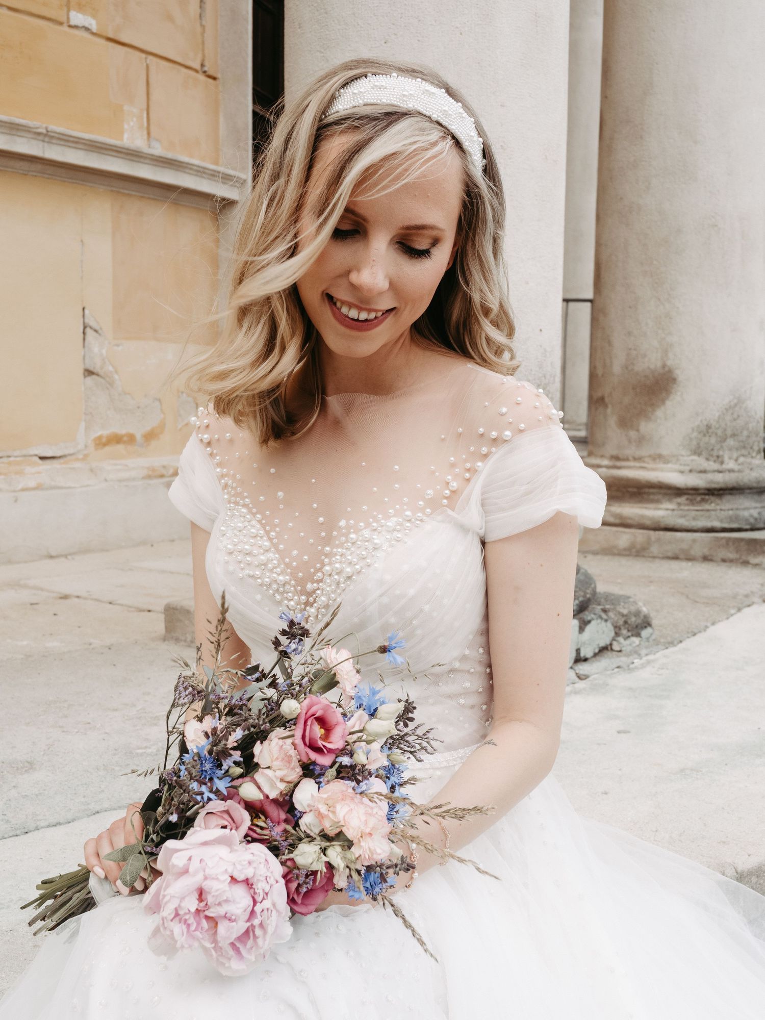 A bride in a lace wedding dress smiles while holding a pink and blue floral bouquet.