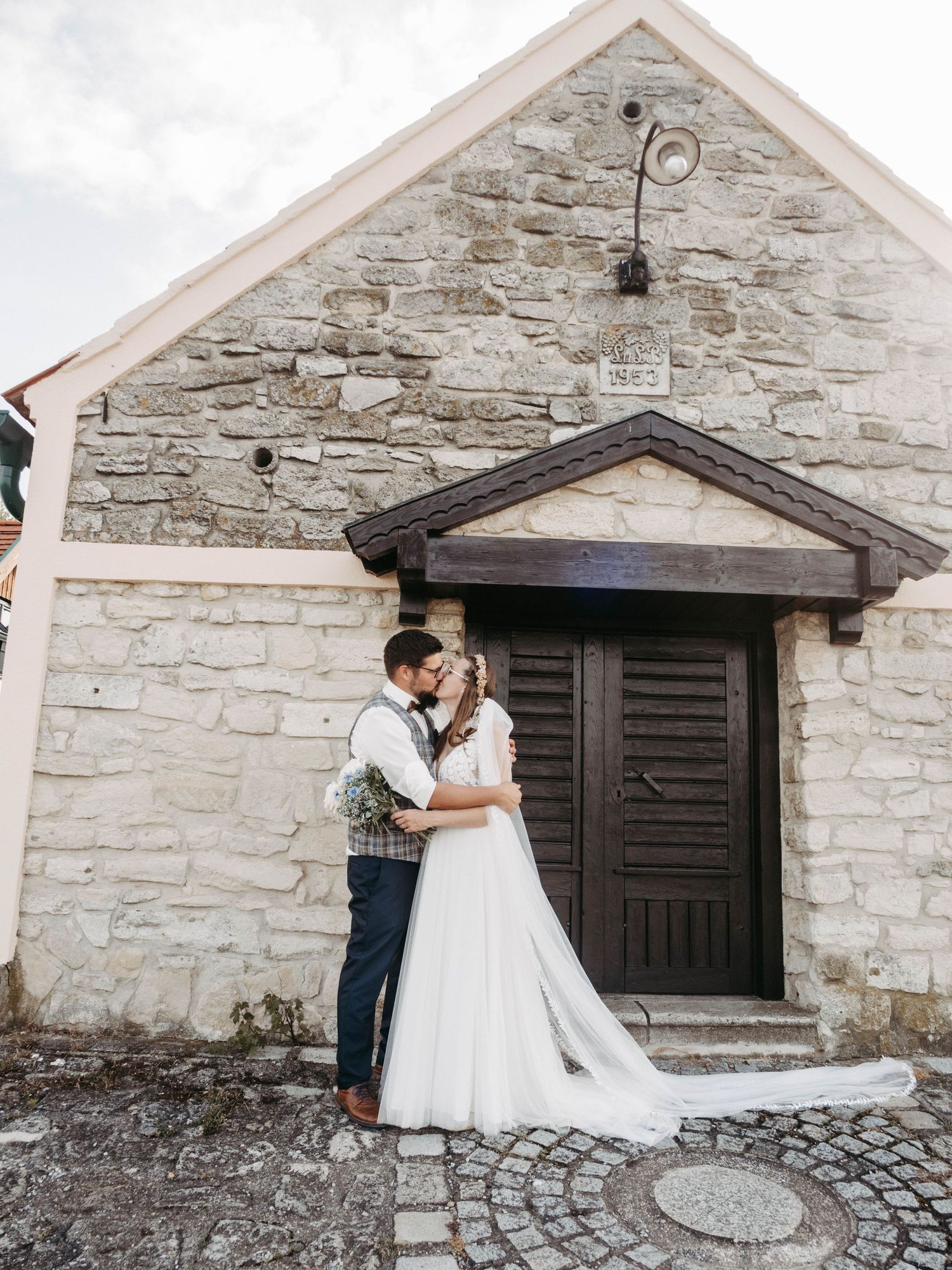Bride and groom share a romantic kiss in front of a rustic stone chapel with a wooden door on their wedding day.
