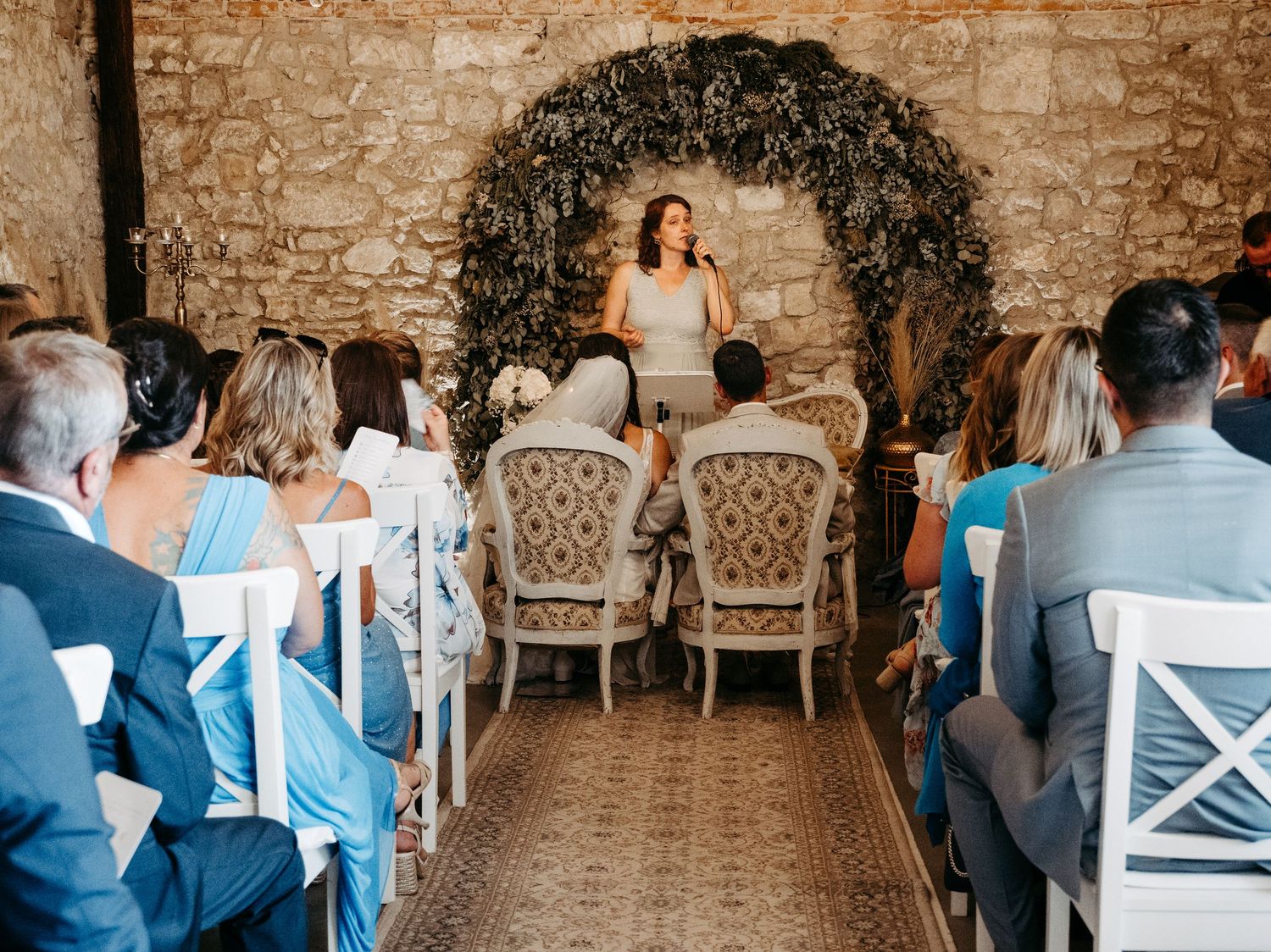 Elegant wedding ceremony in a stone venue with floral arch, white chairs, and officiant addressing seated guests.