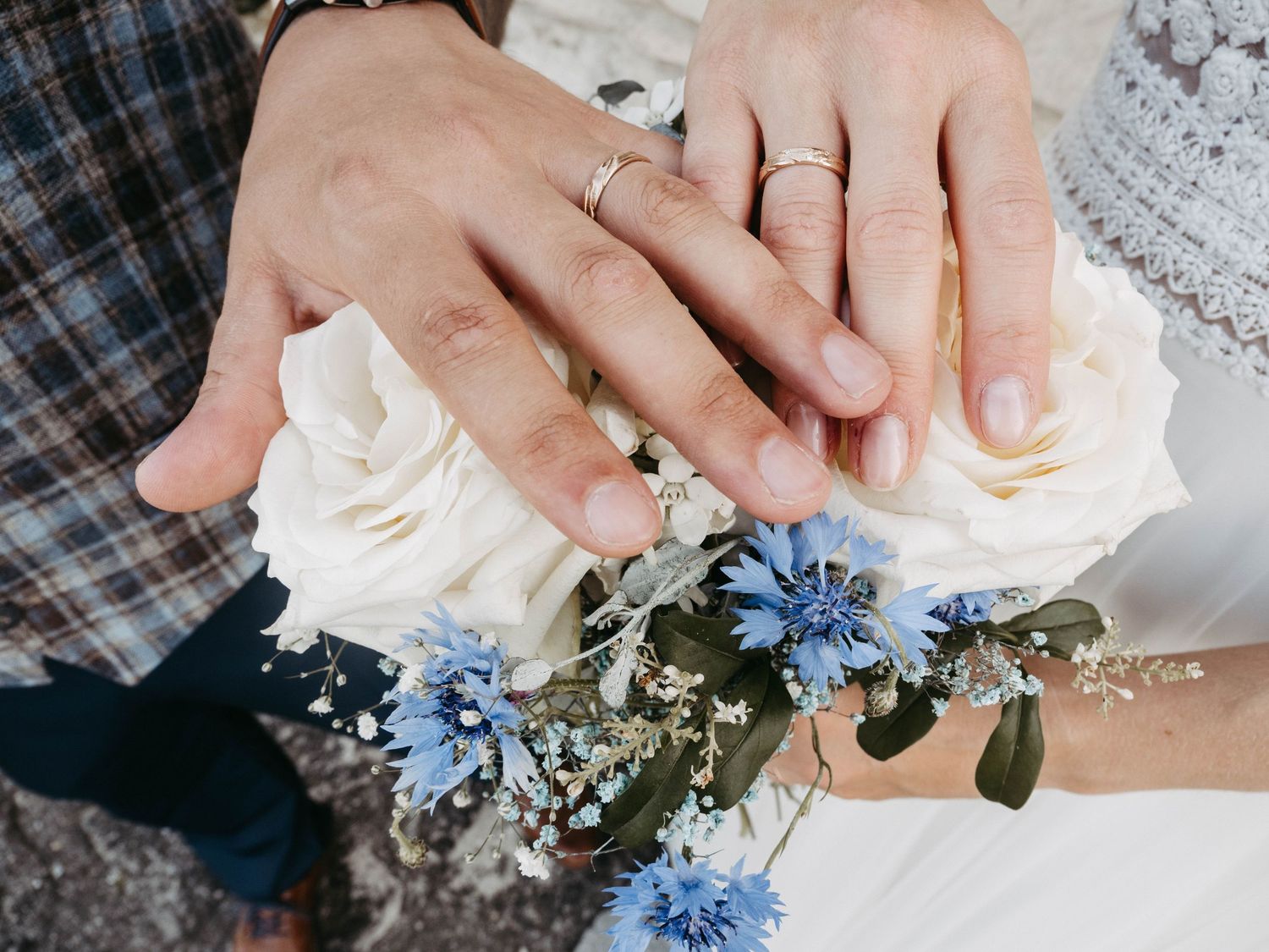 Newlywed couple displaying wedding rings on hands resting over a bridal bouquet of white roses and blue cornflowers.