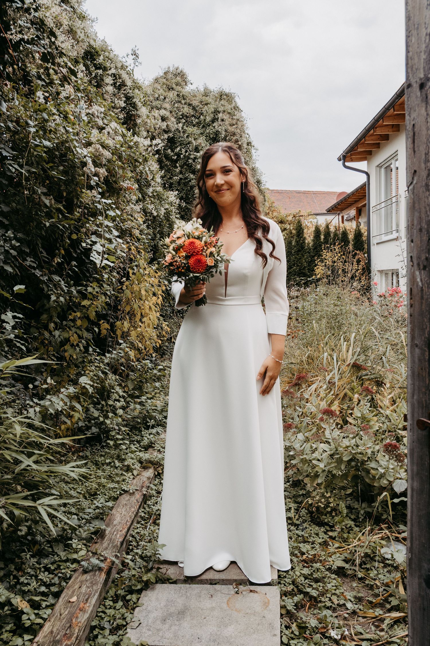 Bride in white long-sleeve gown holding colorful bouquet standing in lush garden pathway.
