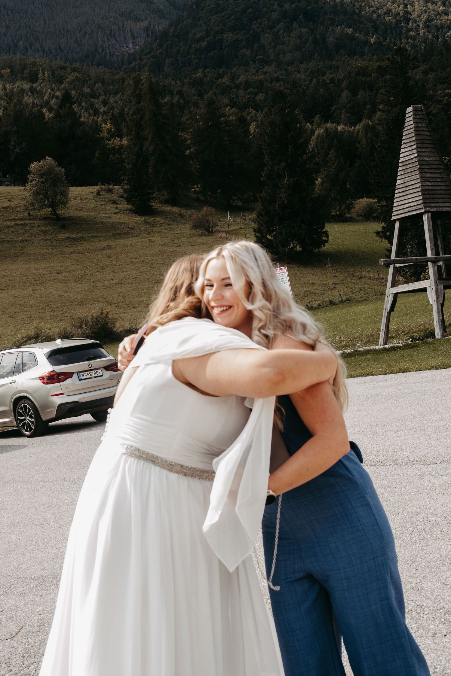 Bride in white gown hugging a guest in blue pants at an outdoor wedding venue with mountain backdrop.
