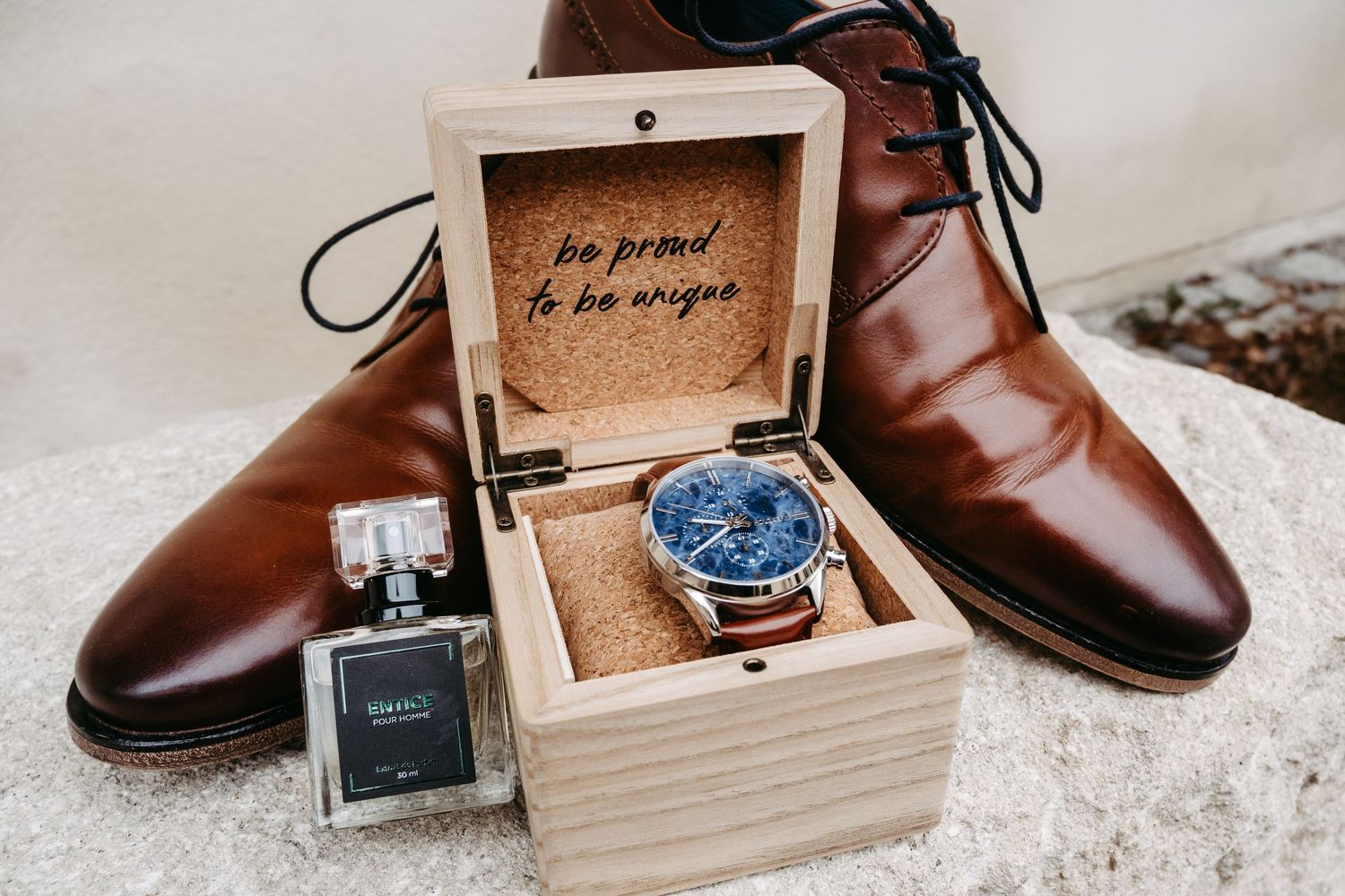 Groom accessories flat lay with blue watch in wooden box, brown leather dress shoes, and cologne bottle.