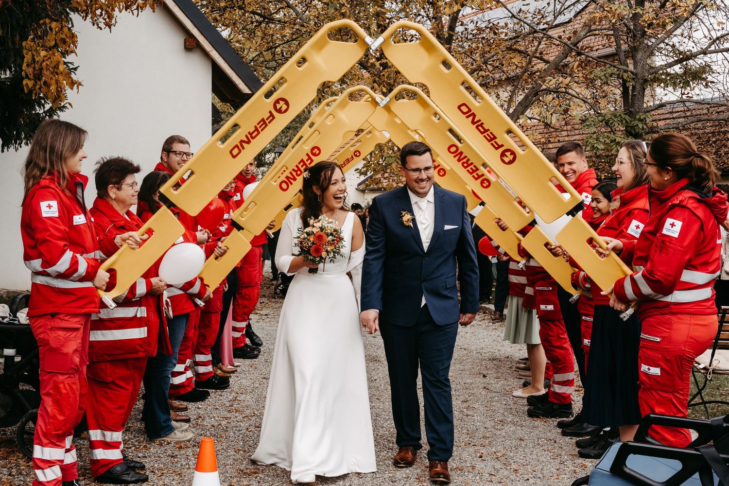 Newlyweds exit ceremony under arch of FERNO stretchers held by Red Cross workers outdoors.