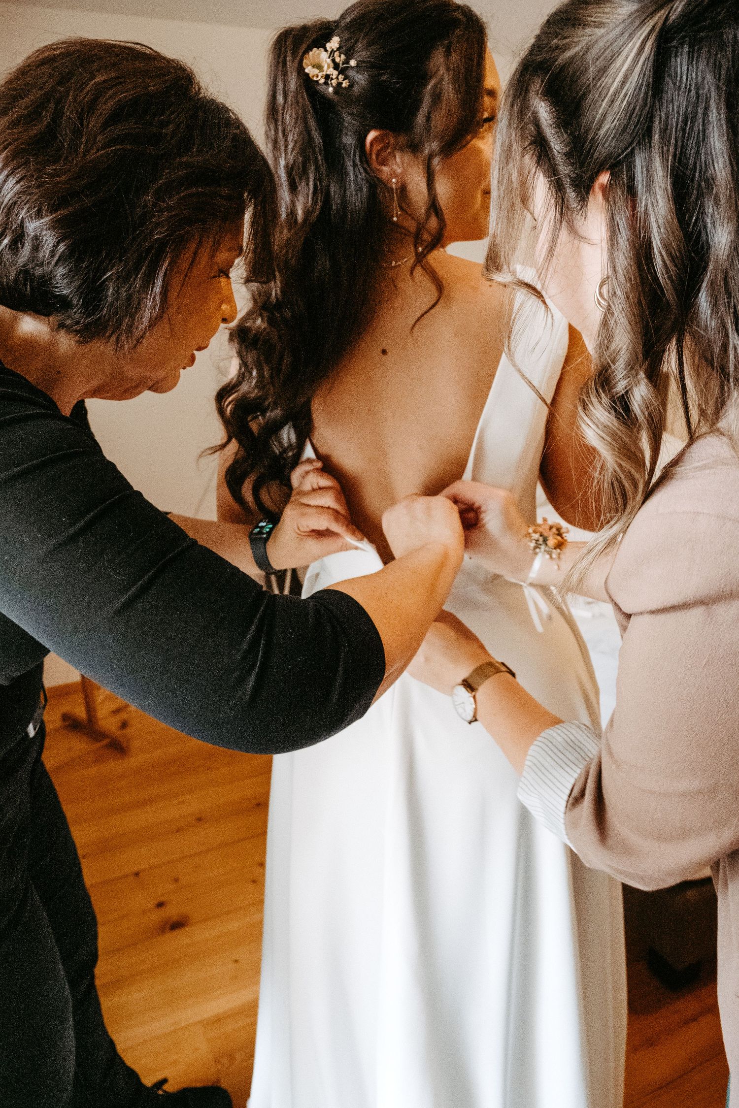 Two women helping bride button up elegant white wedding dress from behind indoors.