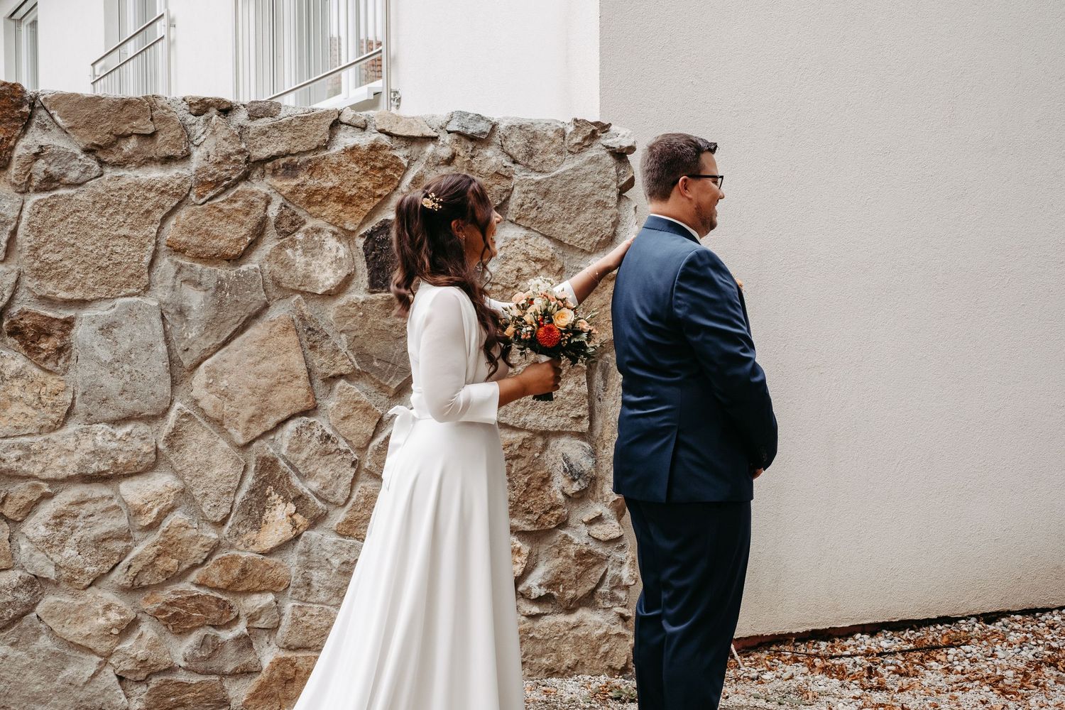 Bride in elegant white gown surprising groom with bouquet during first look ceremony.