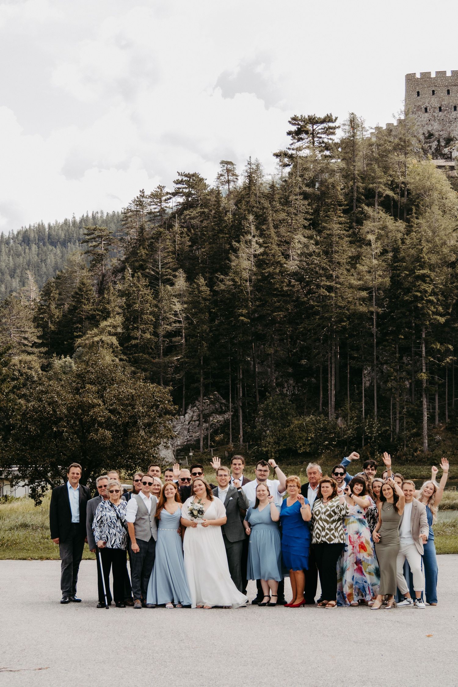 Wedding group photo outdoors with forest and castle backdrop, bride in white gown surrounded by guests.