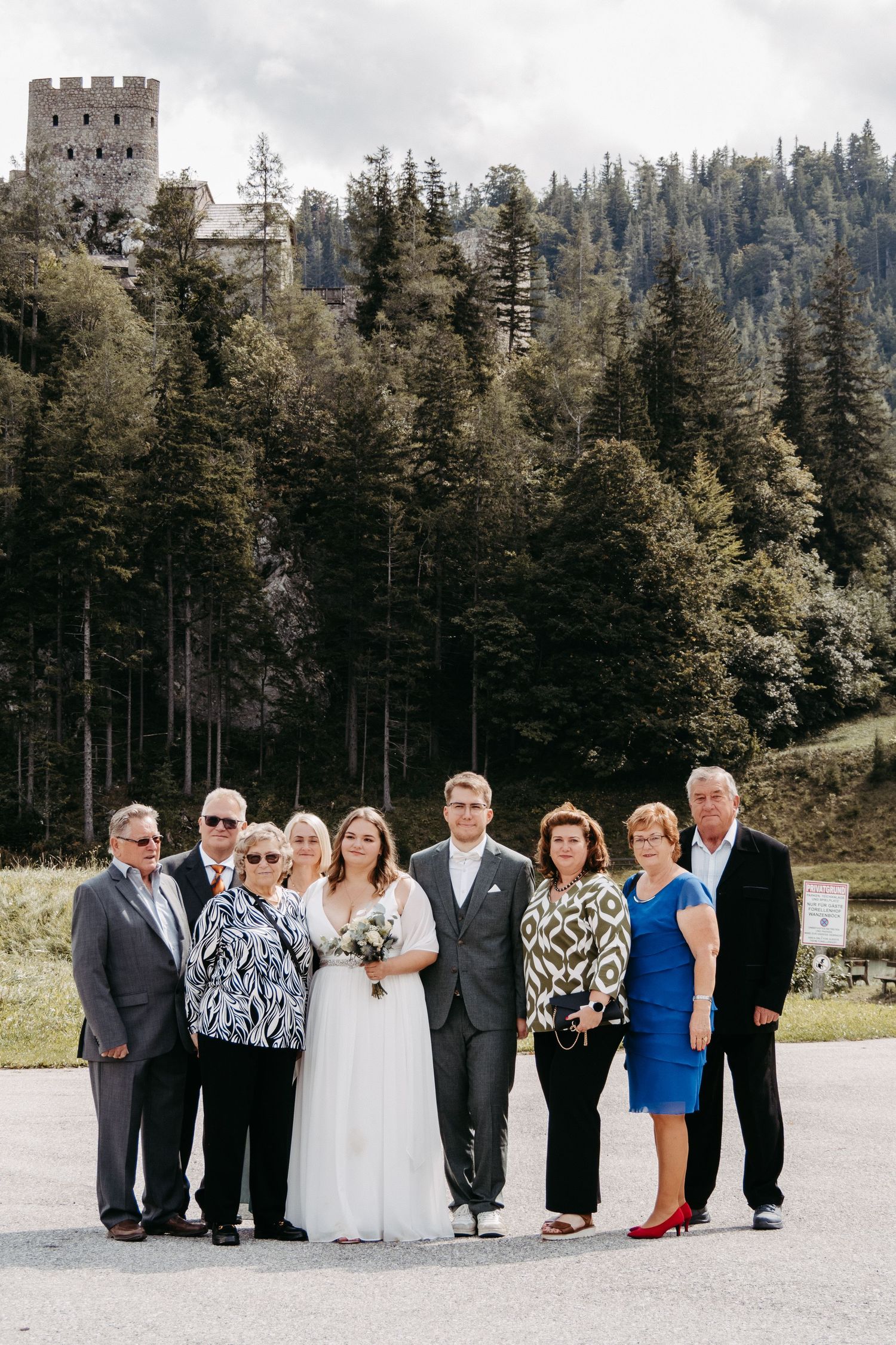 Wedding group photo with bride and groom surrounded by family, set against a forest and castle backdrop.