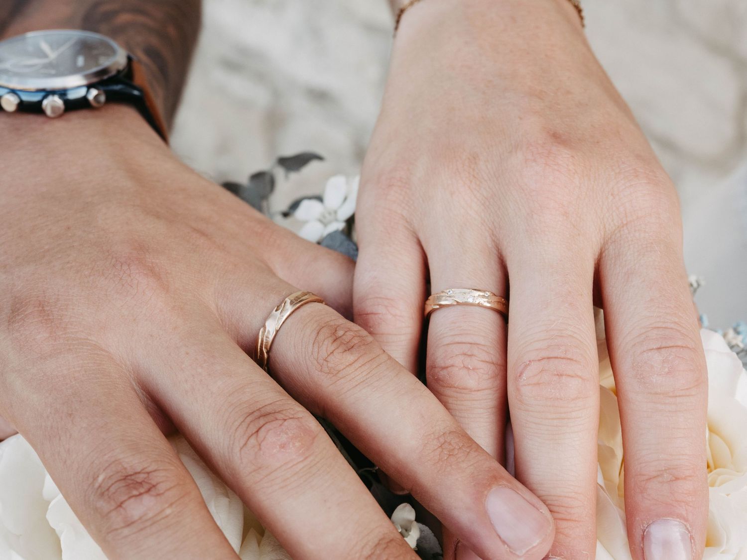 Two hands displaying matching gold wedding bands, one with a watch, symbolizing marriage and commitment.