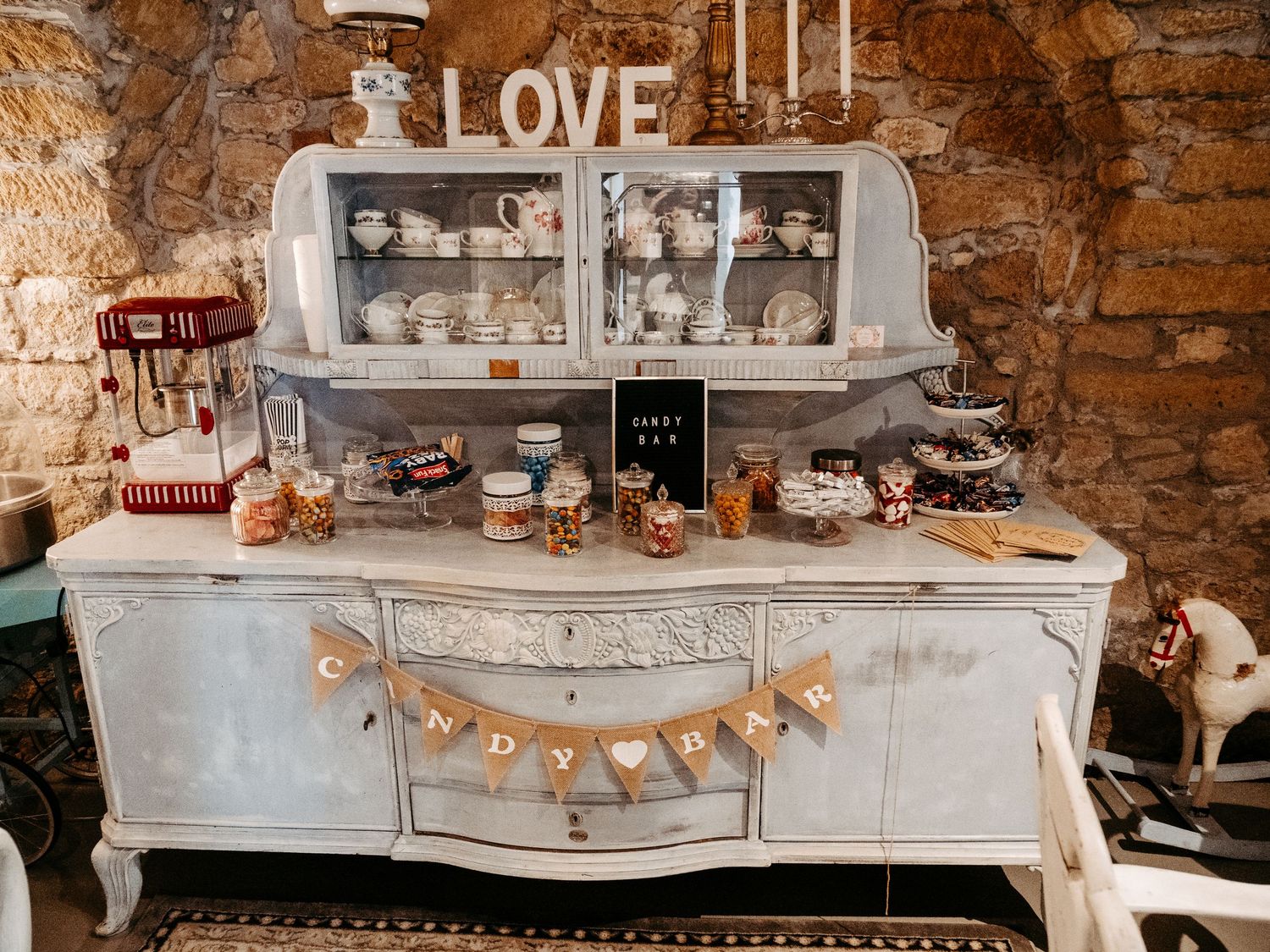 Rustic wedding candy bar display on vintage white dresser with LOVE sign, assorted sweets, and elegant china cabinet backdrop.