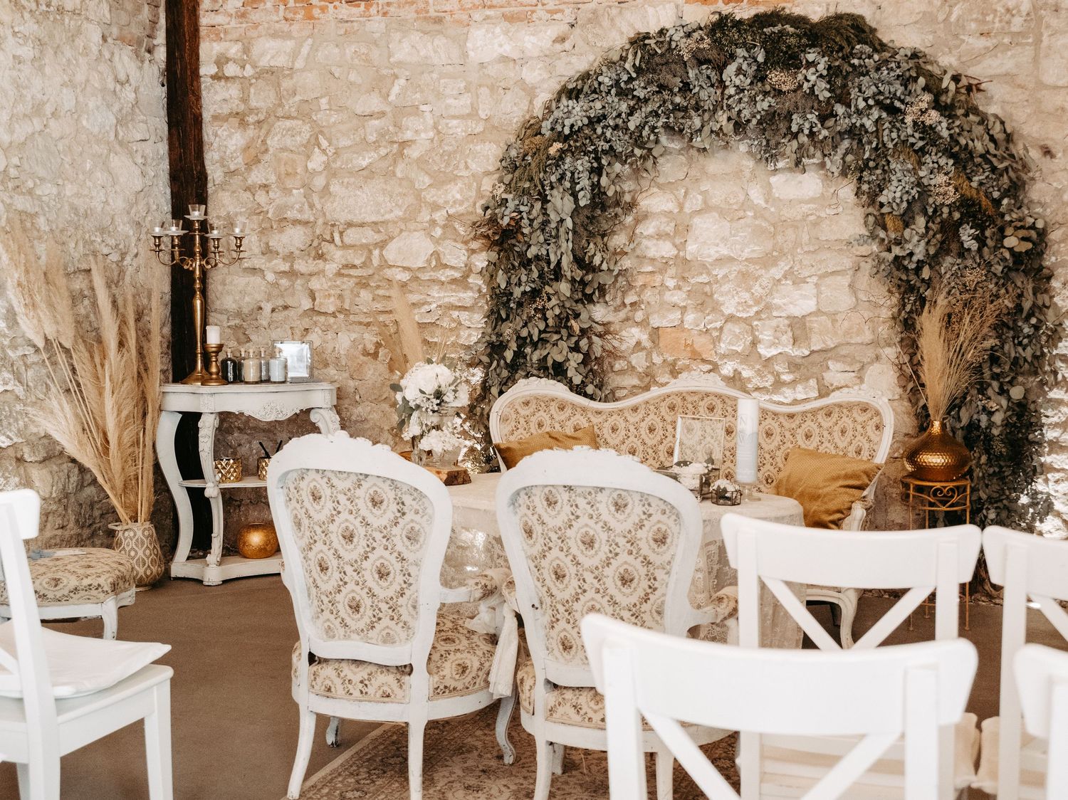 Elegant wedding ceremony setup with floral arch, ornate vintage chairs, and rustic stone wall backdrop.