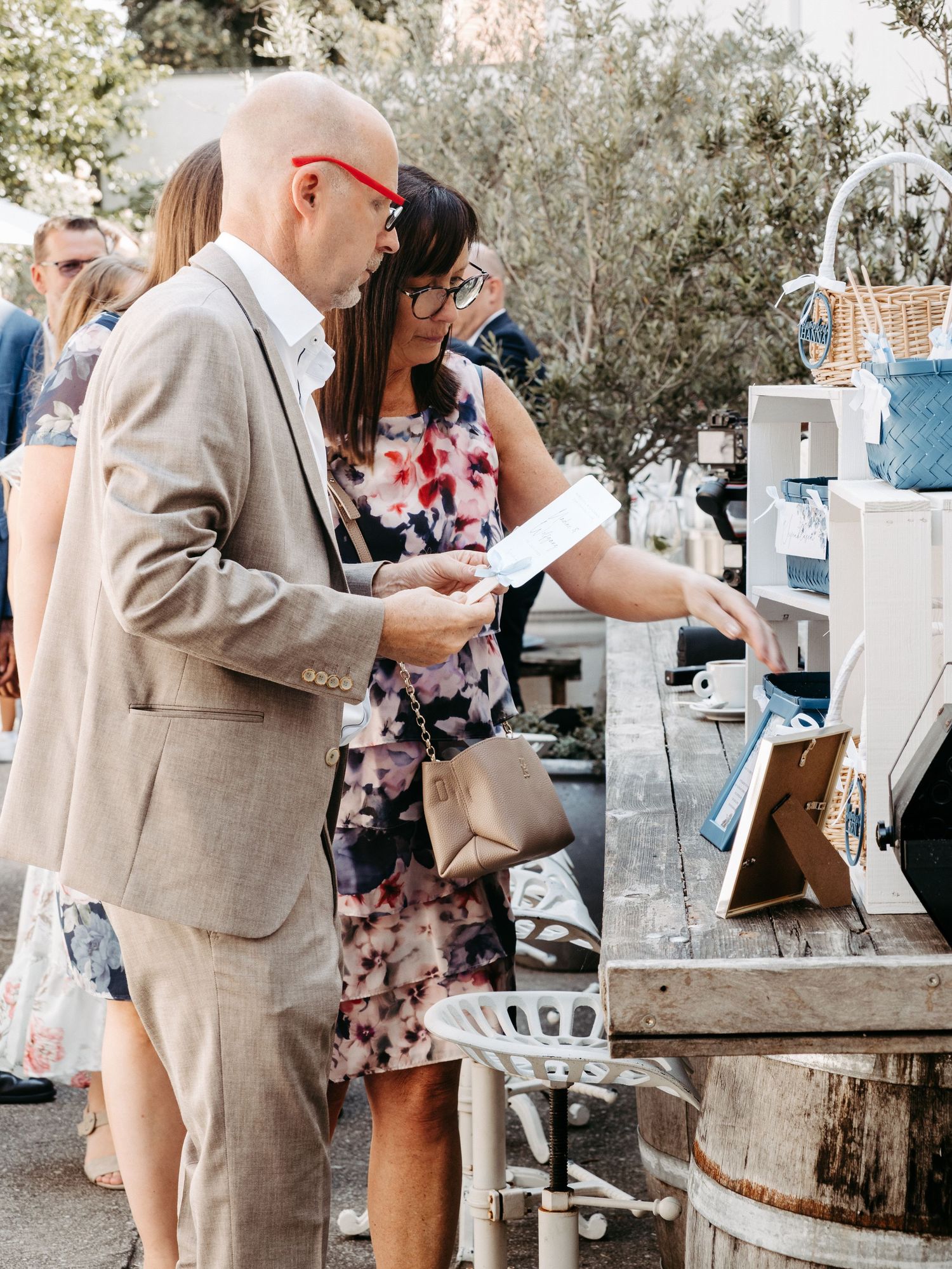 A bald man in a tan suit and woman in floral dress review documents at an outdoor event vendor table.