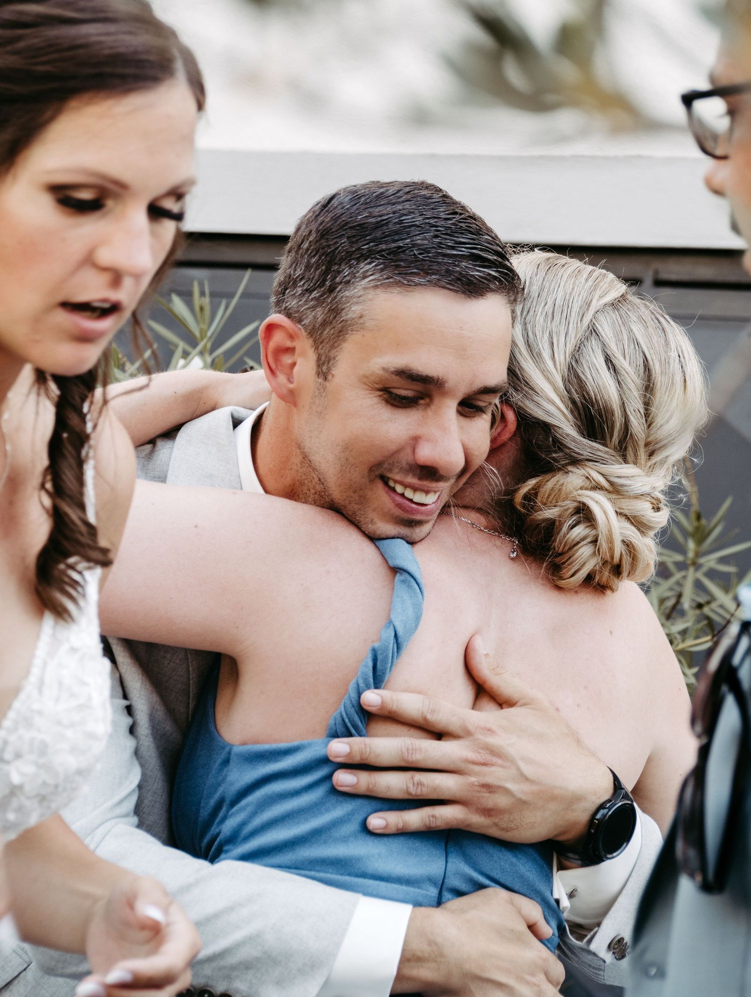 A smiling groom in a gray suit embraces a woman in a blue dress at a wedding celebration outdoors.