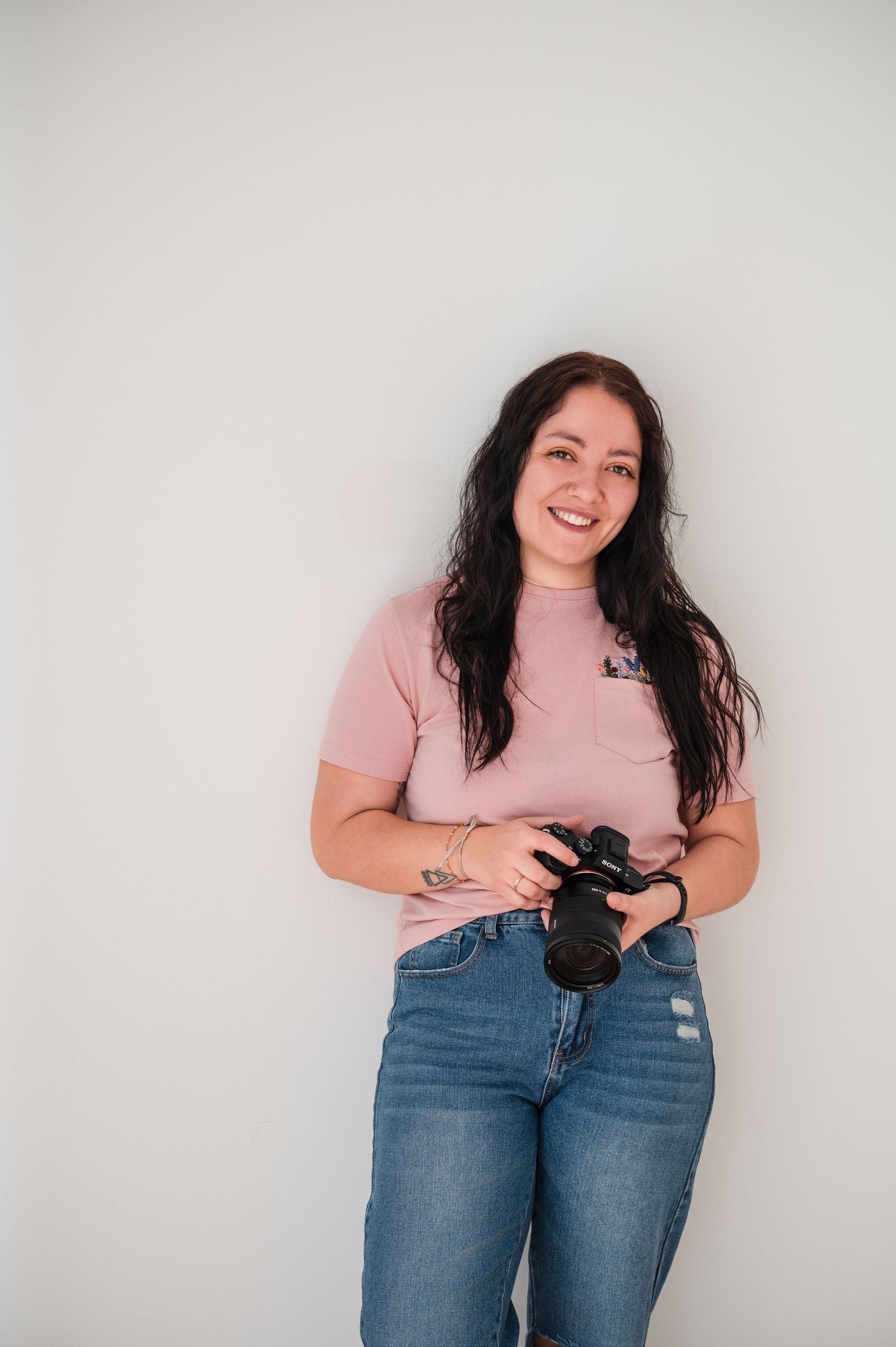 Smiling young woman with dark hair holding a DSLR camera, wearing a pink t-shirt and jeans against a white wall.