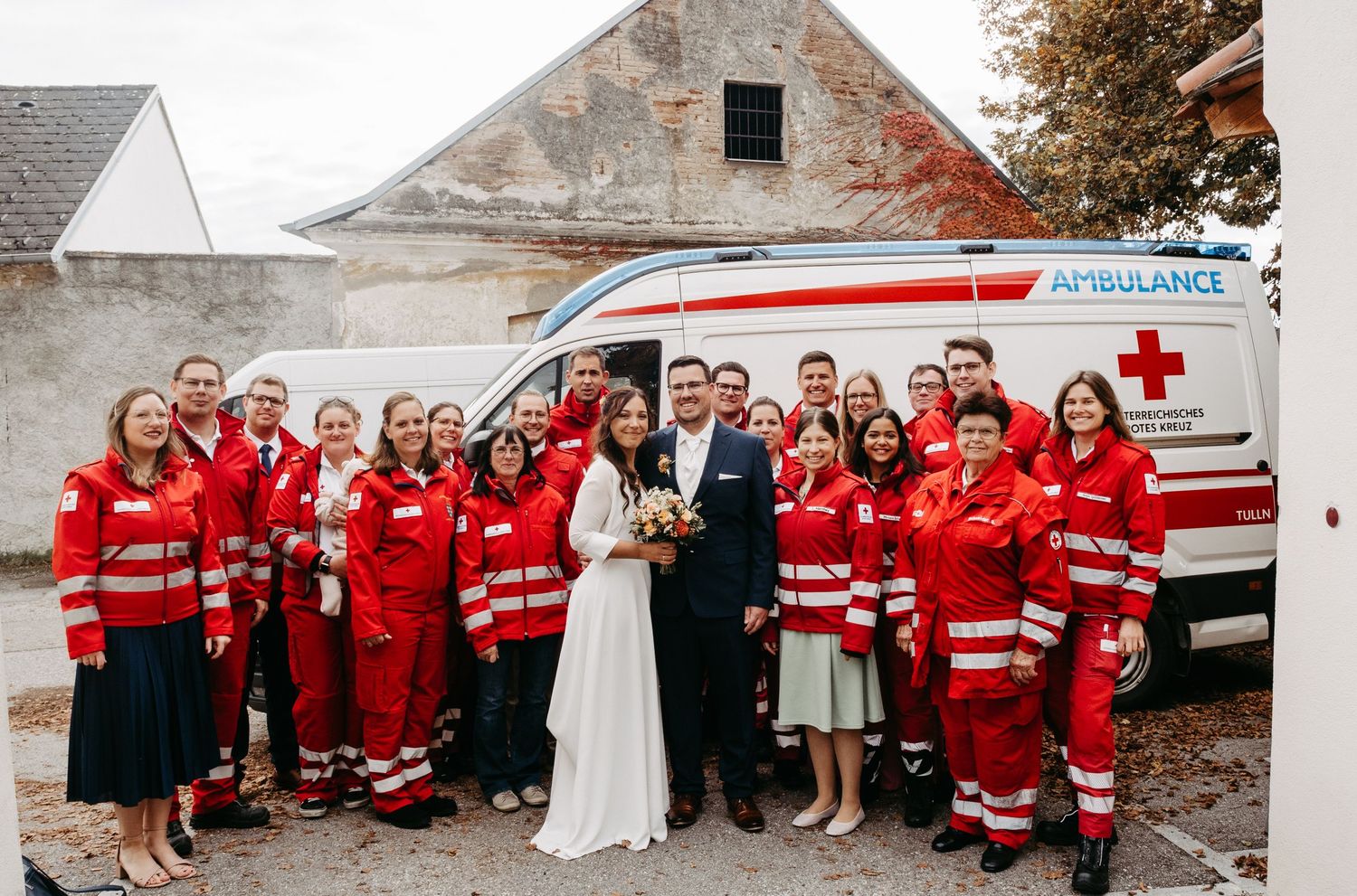 Wedding couple posing with Red Cross ambulance volunteers in red uniforms at rustic outdoor venue.