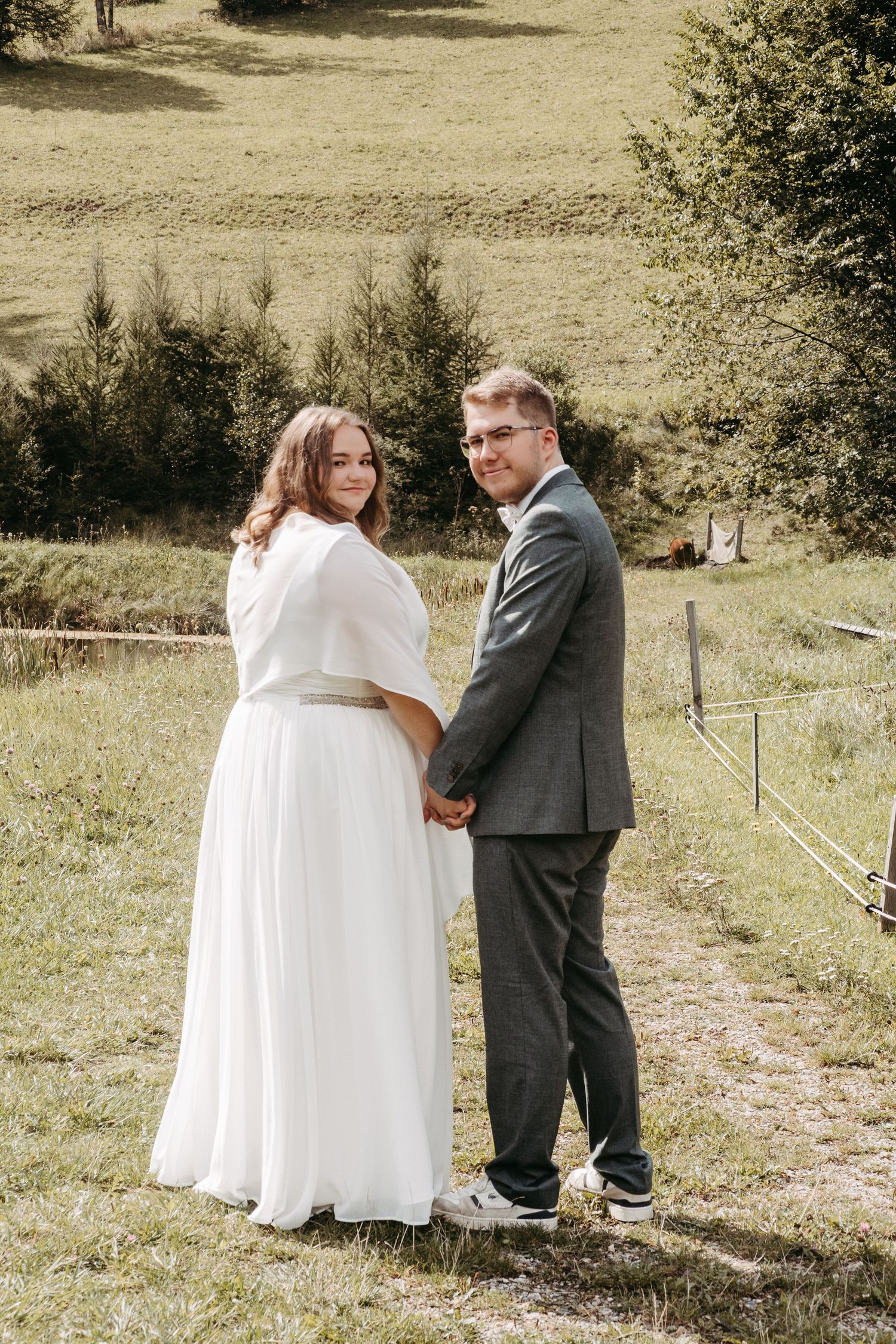 Bride in white dress and groom in gray suit holding hands outdoors on green hillside for wedding portrait.