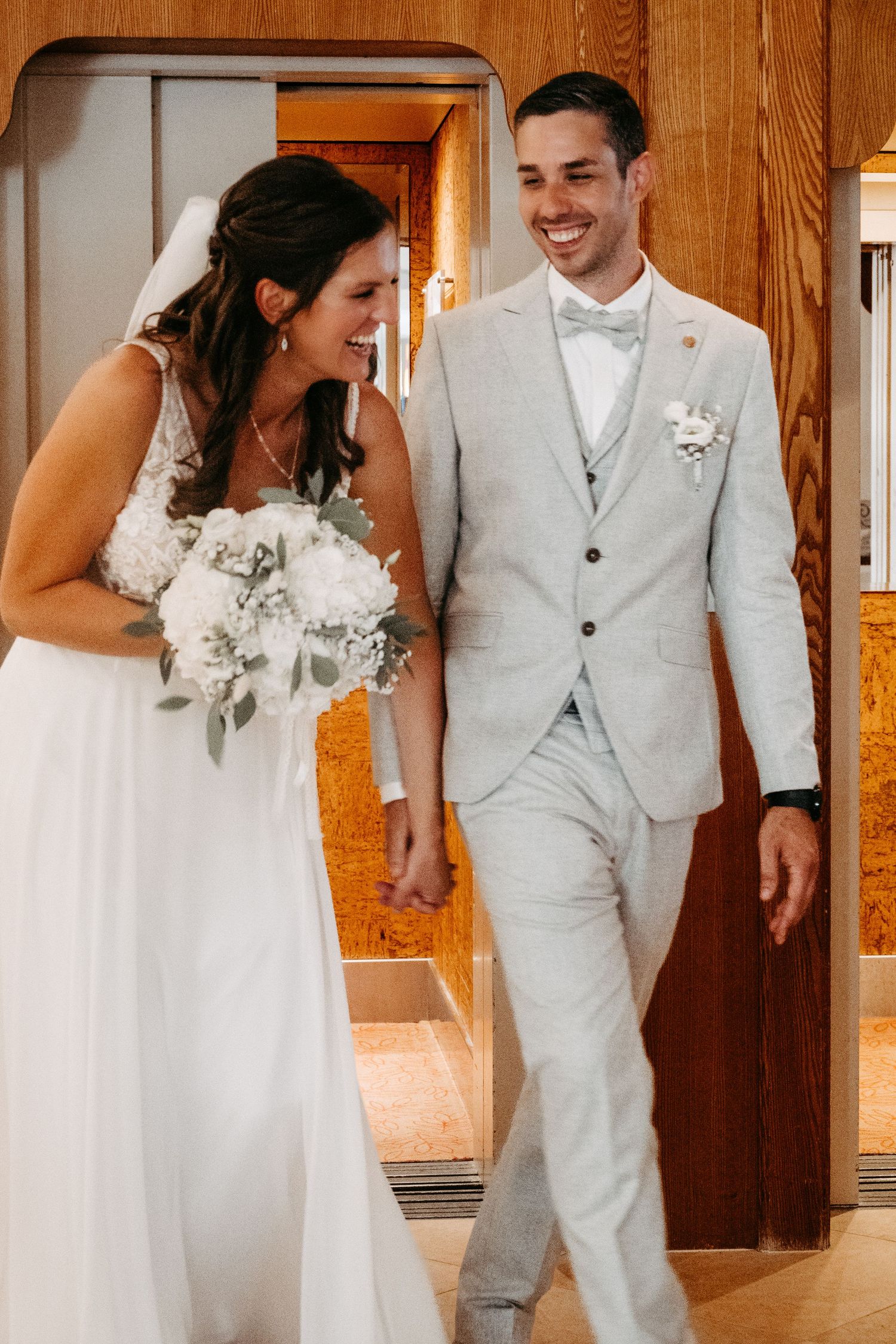 Smiling bride in white gown holding bouquet walks hand-in-hand with groom in light gray suit inside wedding venue.