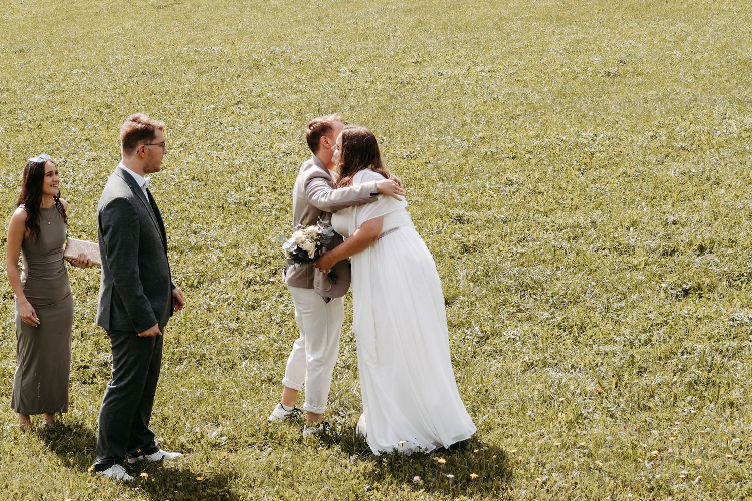 Two brides share a kiss during an outdoor wedding ceremony on a green grass field with guests watching nearby.