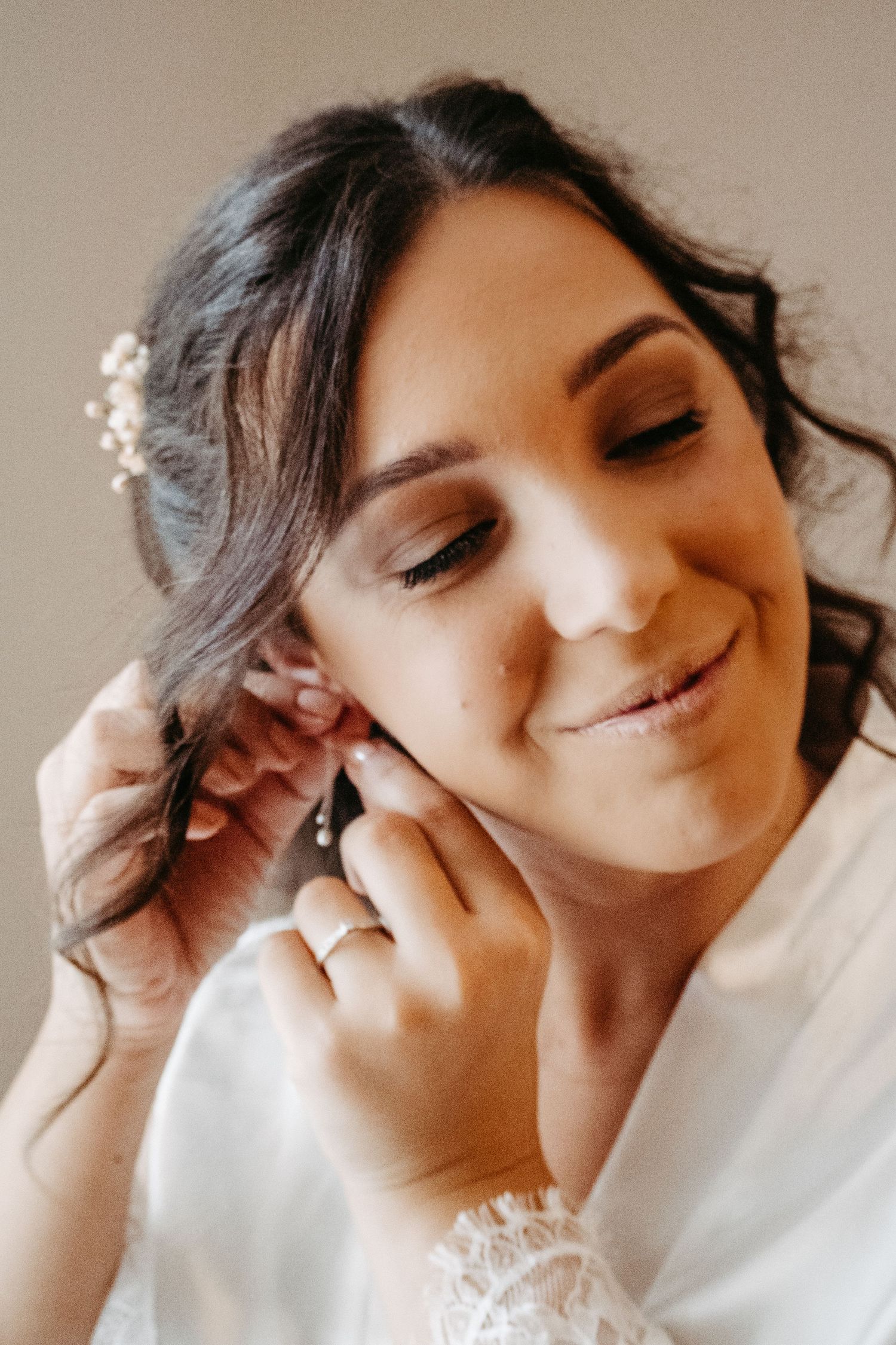 Bride smiling while putting on earrings, wearing white robe with floral hair accessory on wedding day.