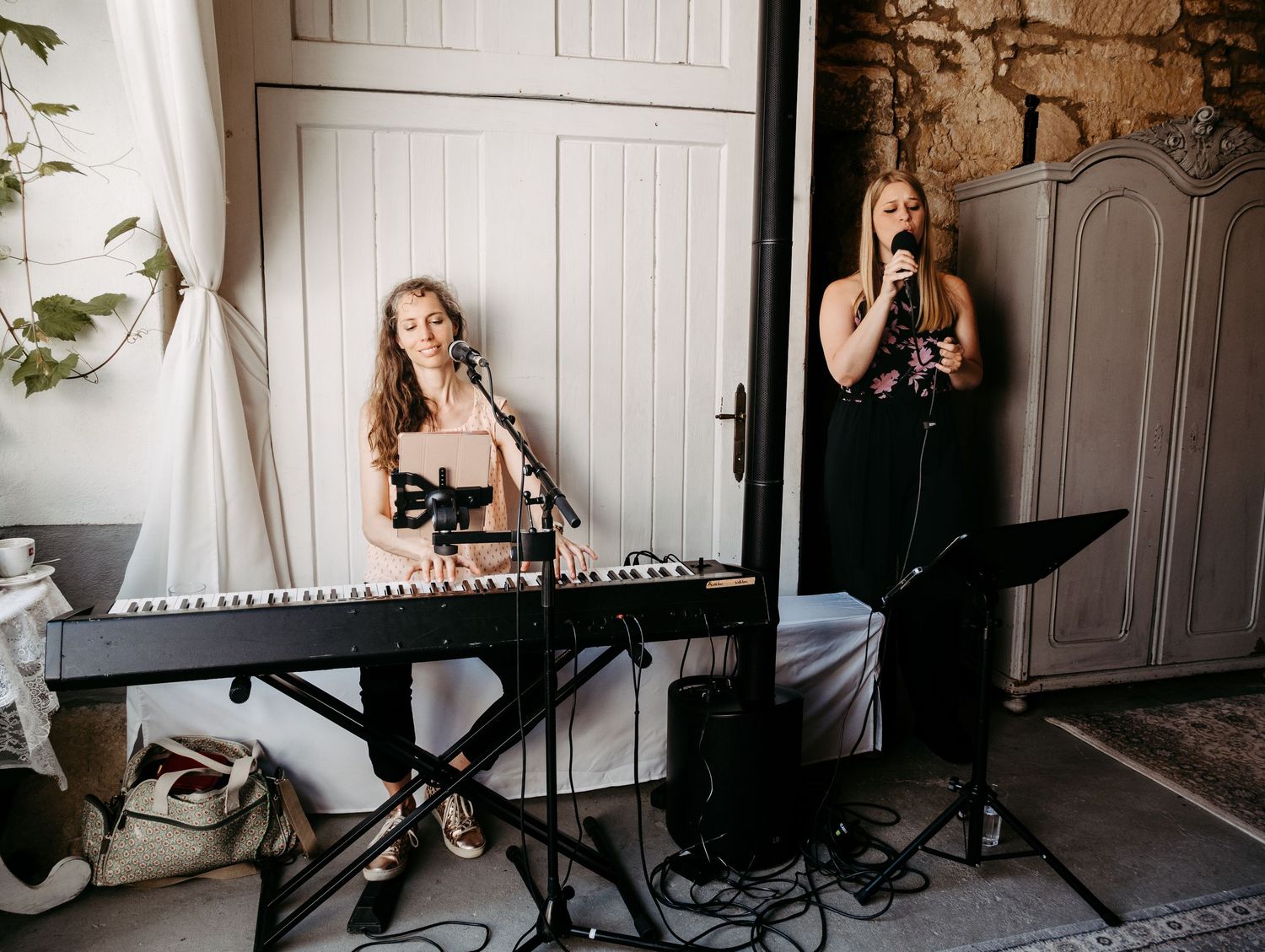 Two women performing live music, one playing keyboard and one singing into a microphone in a rustic venue.