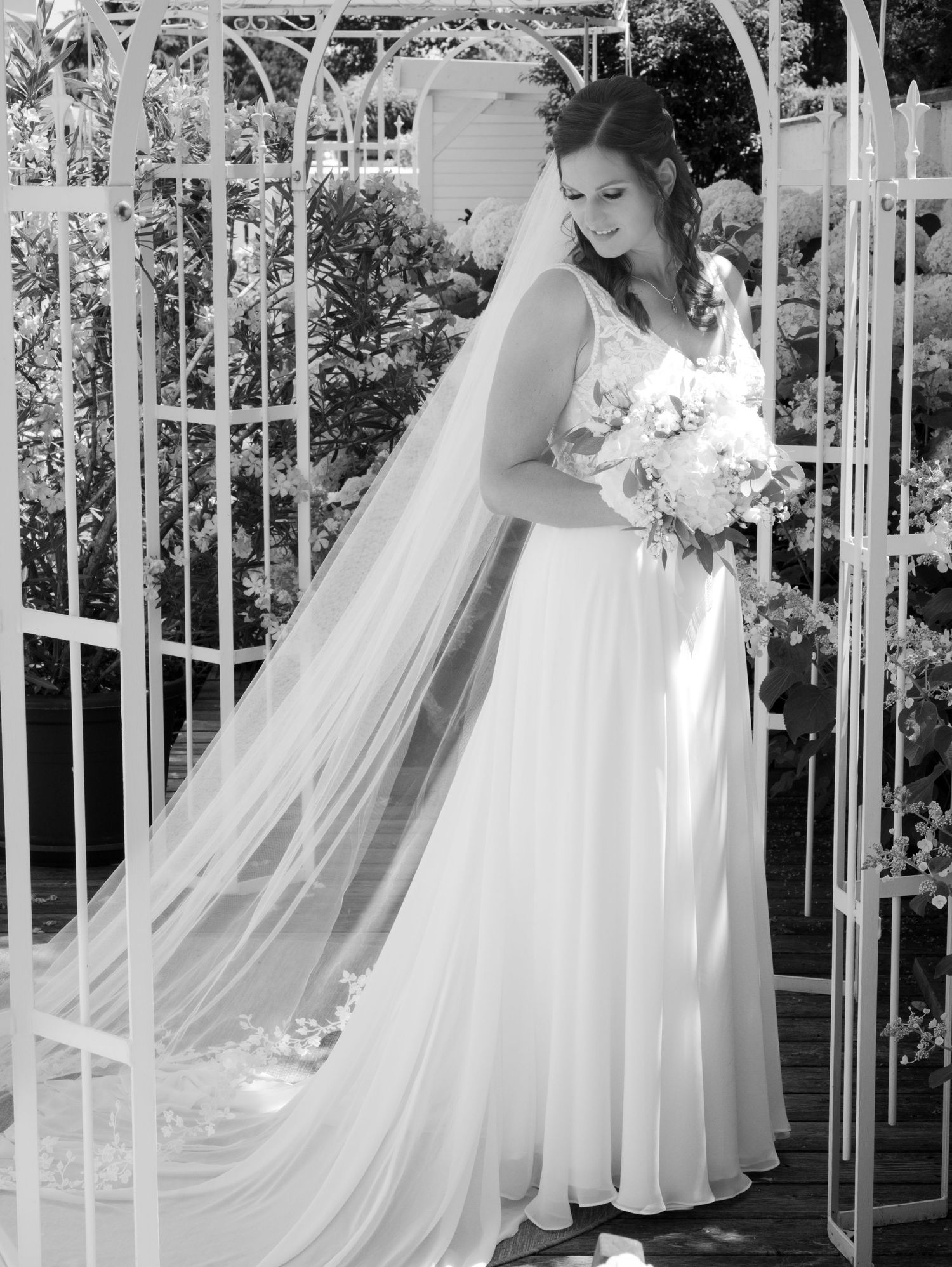 Bride in a flowing white gown and long veil holds a bouquet inside a white garden gazebo, black and white photo.