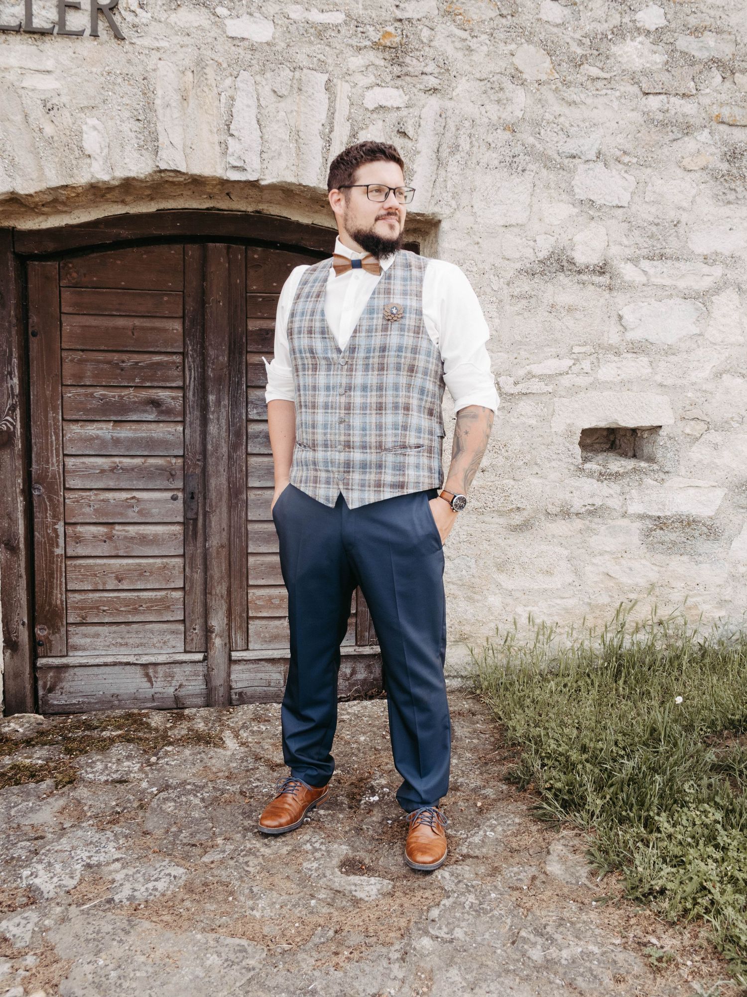 Man in plaid vest, bow tie, blue trousers and tan shoes posing by rustic stone wall and wooden door.