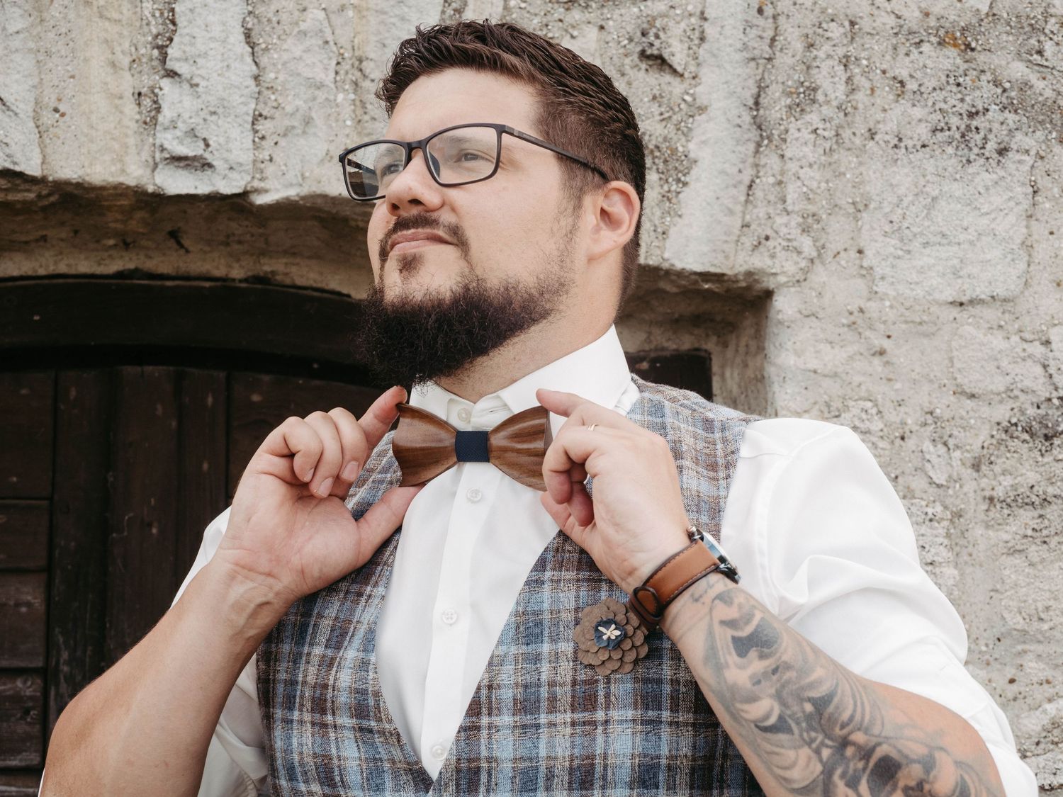 Tattooed man in plaid vest and white shirt adjusting brown bow tie, wearing glasses outdoors.