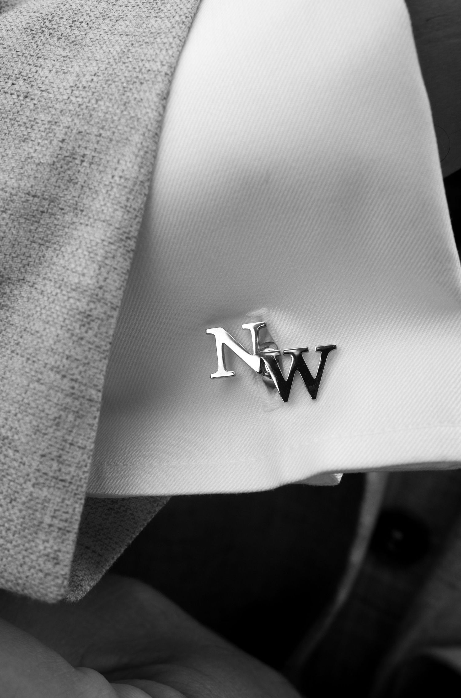 Black and white close-up of silver NW monogram cufflink on white dress shirt sleeve with gray suit jacket.