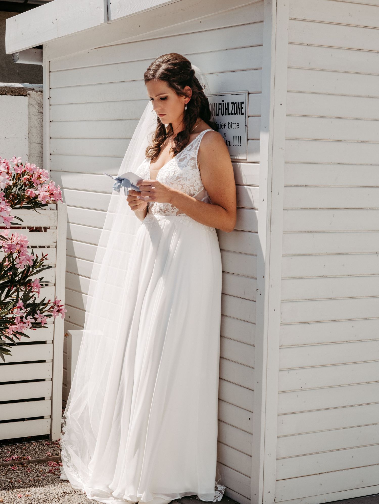 Bride in white lace wedding gown and veil reads a note while leaning against a white wooden building outdoors.
